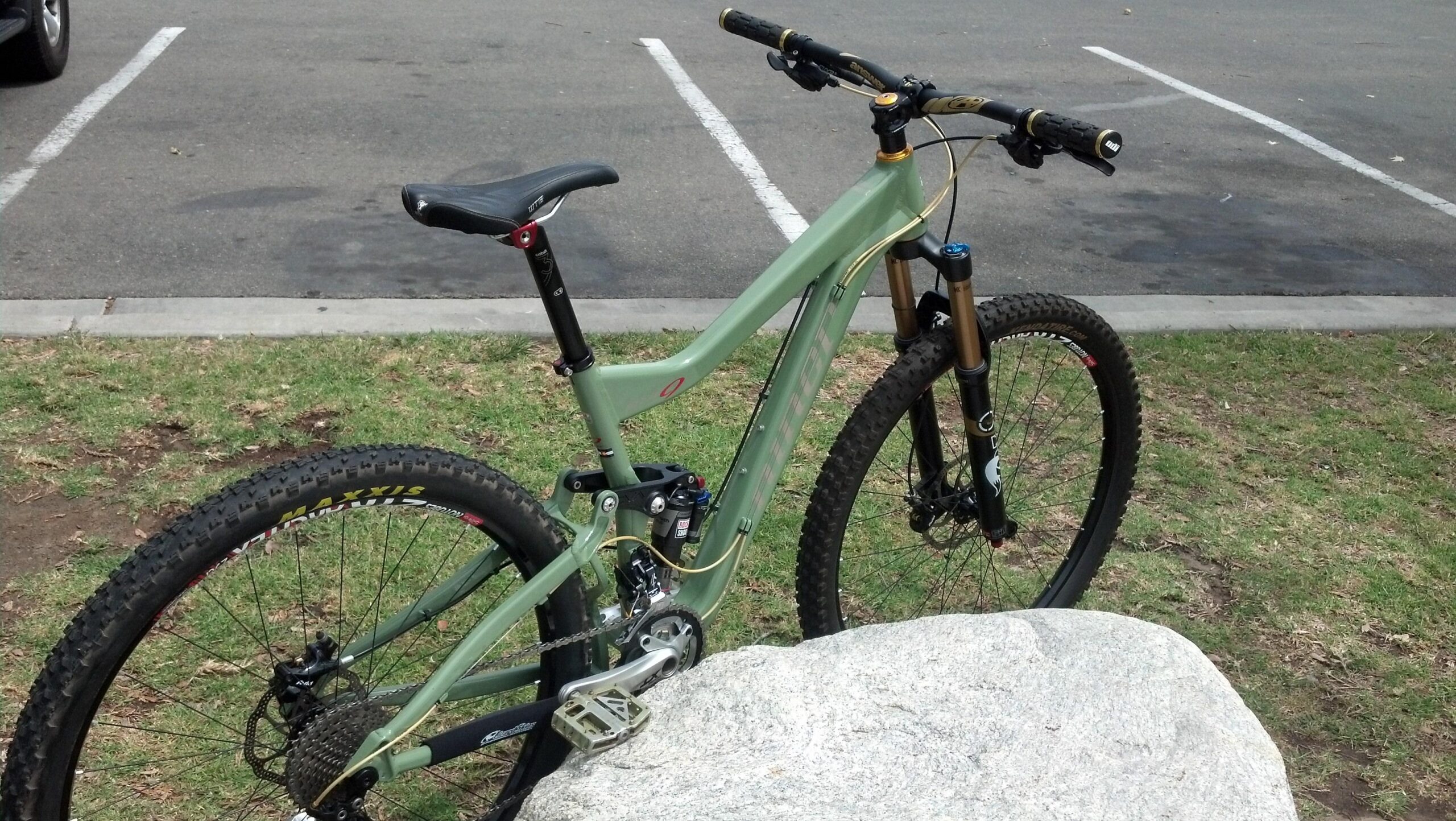 Niner W.F.O. 9: A mountain bike leaning against a rock in an outdoor parking area. The bike features a light green frame, thick tires with tread patterns, and adjustable handlebars. In the background, there are parking lines and patches of grass, indicating a recreational setting.