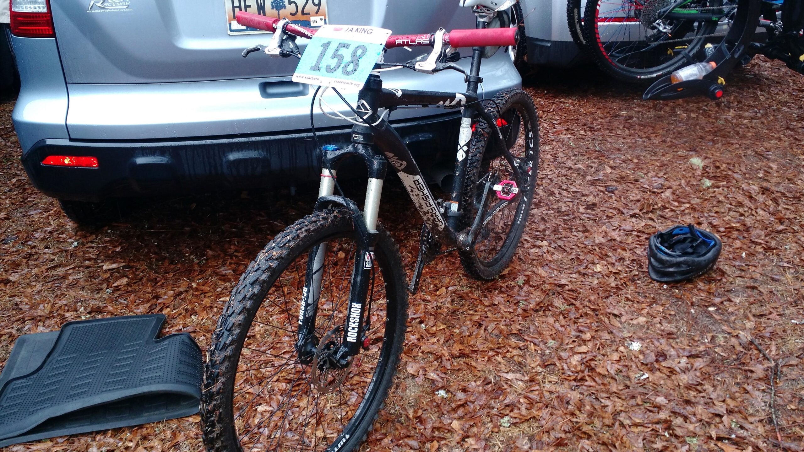 Gary Fisher Wahoo: A mountain bike with number 158 attached to the handlebars, parked on a bed of fallen leaves near a vehicle. The bike features a shiny frame, muddy tires, and a suspension fork, with a helmet lying on the ground nearby.