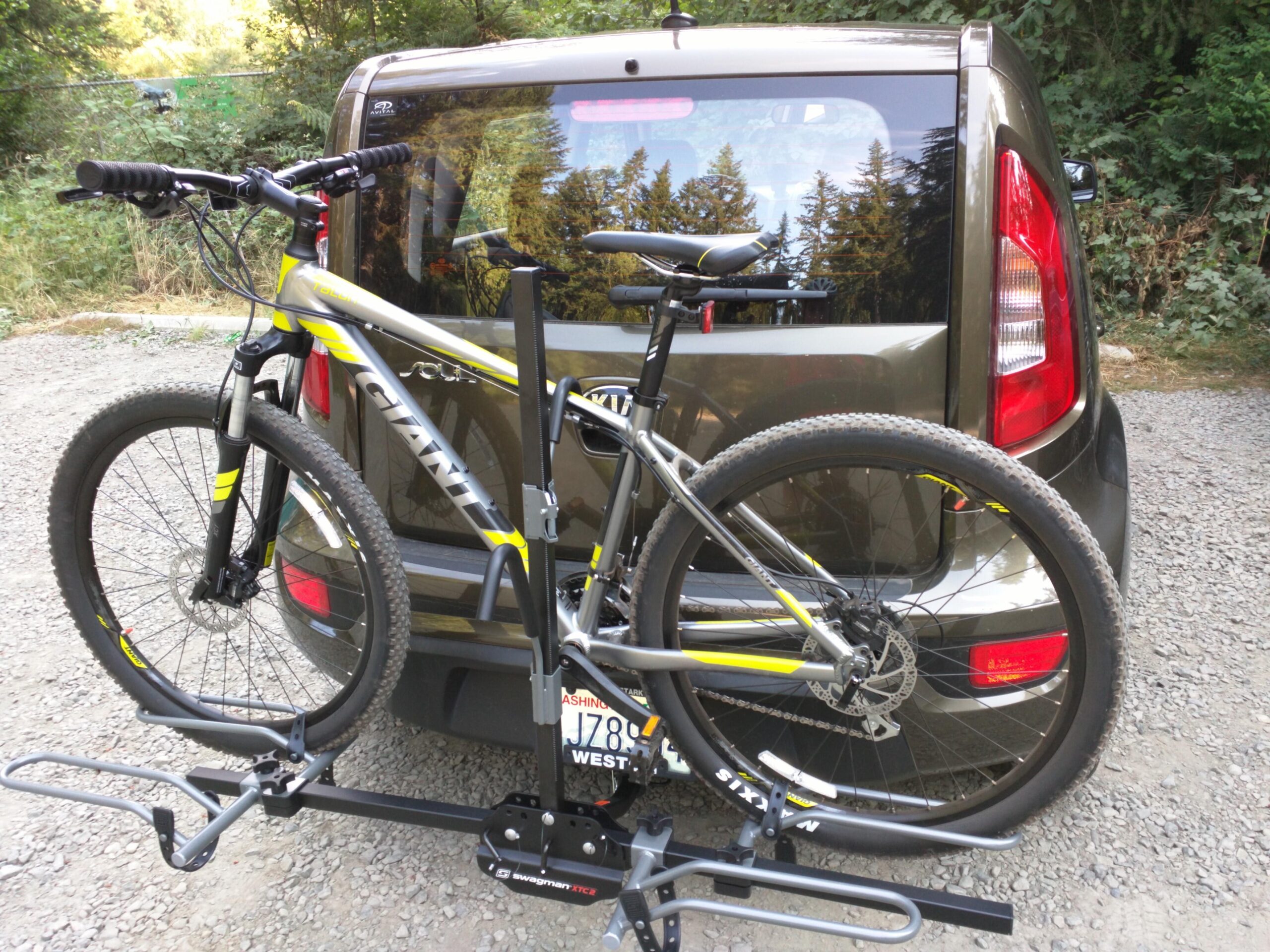 Giant Talon 27.5 4: A mountain bike secured on a bike rack attached to the rear of a vehicle, parked on a gravel surface with trees in the background. The bike features bright yellow accents and is designed for off-road cycling. The vehicle is compact and has a dark metallic color.