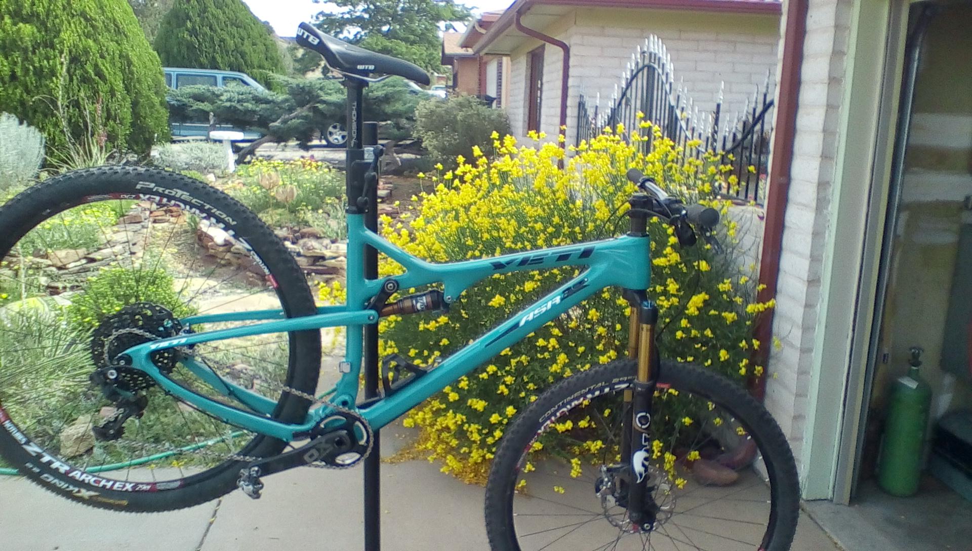 Yeti ASR 5 Carbon: A turquoise mountain bike mounted on a repair stand, featuring a dual suspension design and disc brakes. In the background, a vibrant bush with yellow flowers and greenery surrounds a residential area. There are also pebbled landscaping features and a visible vehicle parked in the distance.