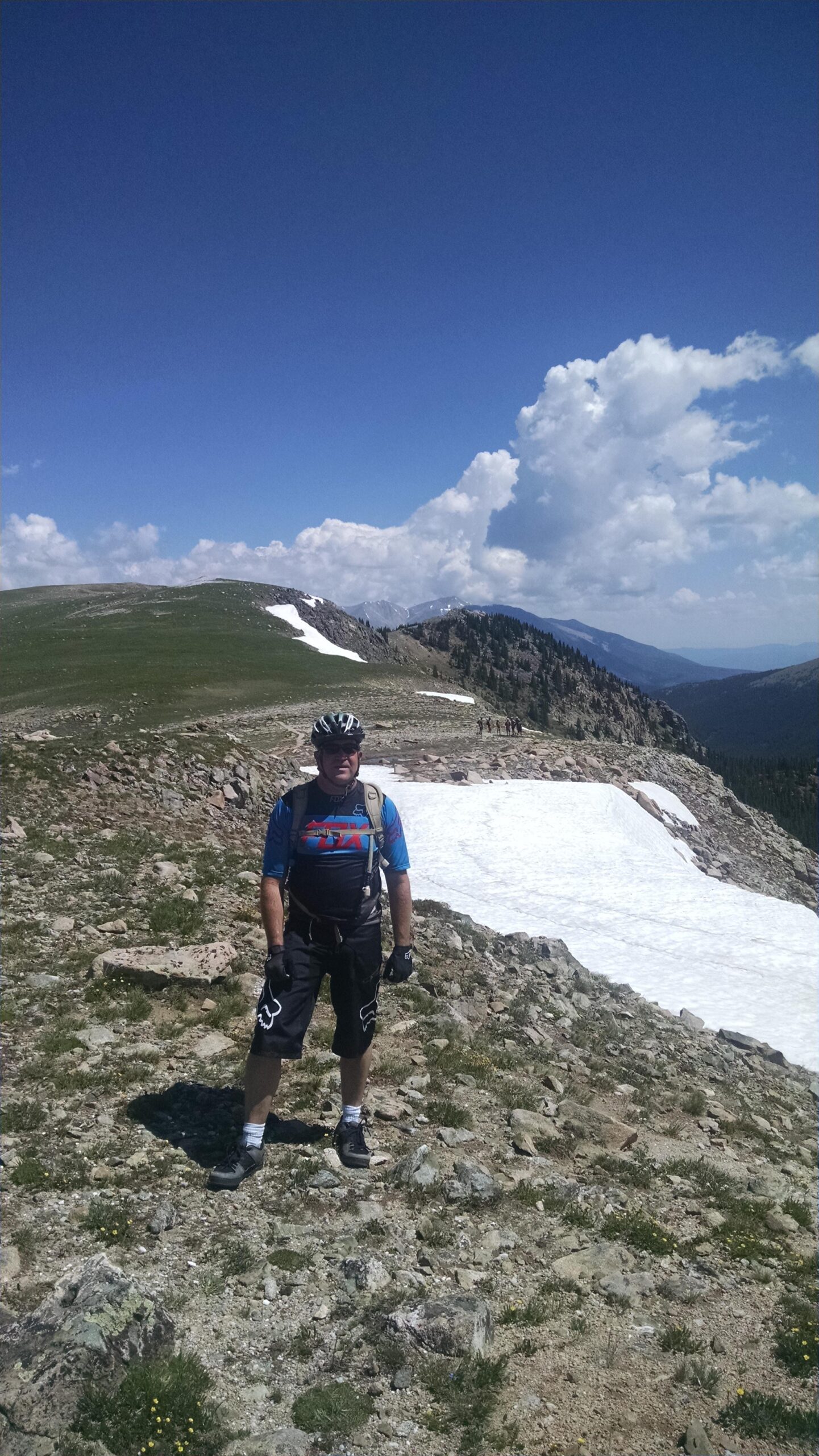 A person stands on a rocky mountain trail with patches of snow and lush green vegetation around. They are wearing cycling gear, including a helmet and gloves, and are positioned against a backdrop of clear blue skies and distant mountain peaks. The scene captures the essence of outdoor adventure and mountain biking. Monarch Crest Trail mountain bike trail.