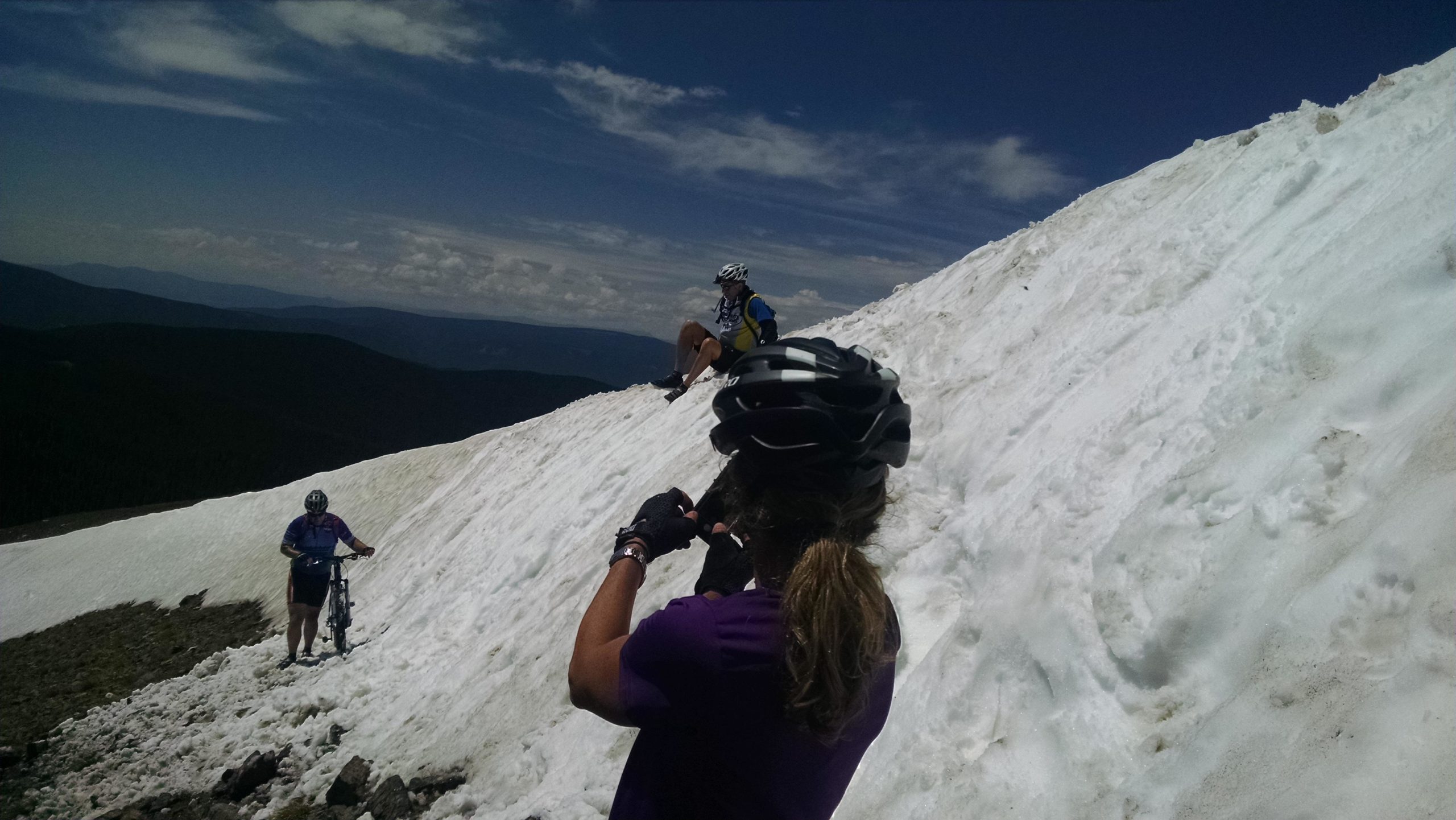 Two mountain bikers are enjoying a snowy landscape at high altitude. One biker stands near their bicycle, while another sits on the snow-covered slope, both wearing helmets and outdoor clothing. The backdrop features a mix of mountains and a cloudy sky. Monarch Crest Trail mountain bike trail.