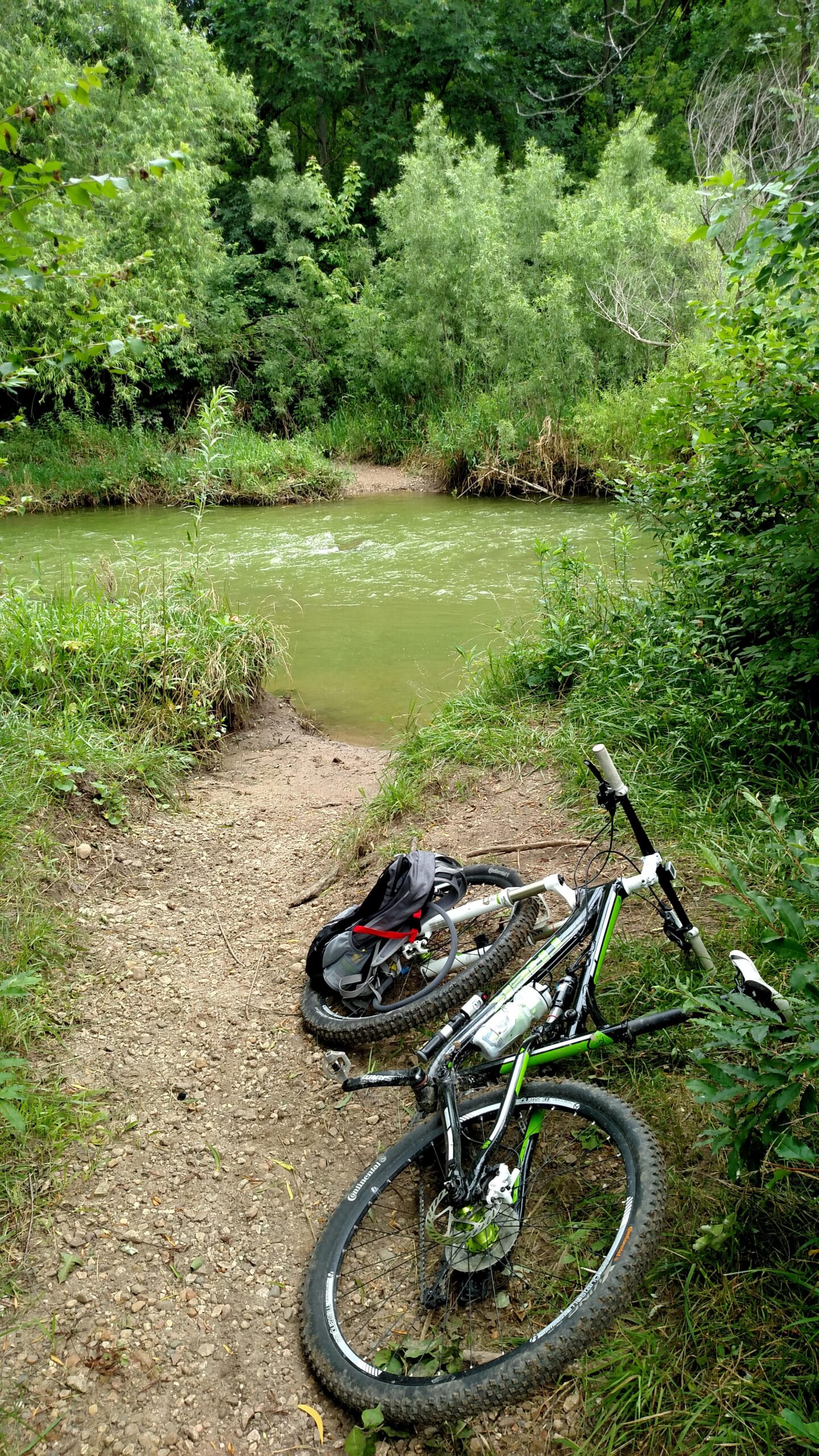 Trek Superfly 100 AL: A mountain bike rests on a gravel path beside a calm green river, surrounded by lush greenery and trees. A small backpack is placed next to the bike, with part of the bank visible in the background. The scene conveys a peaceful outdoor setting ideal for biking and exploration.