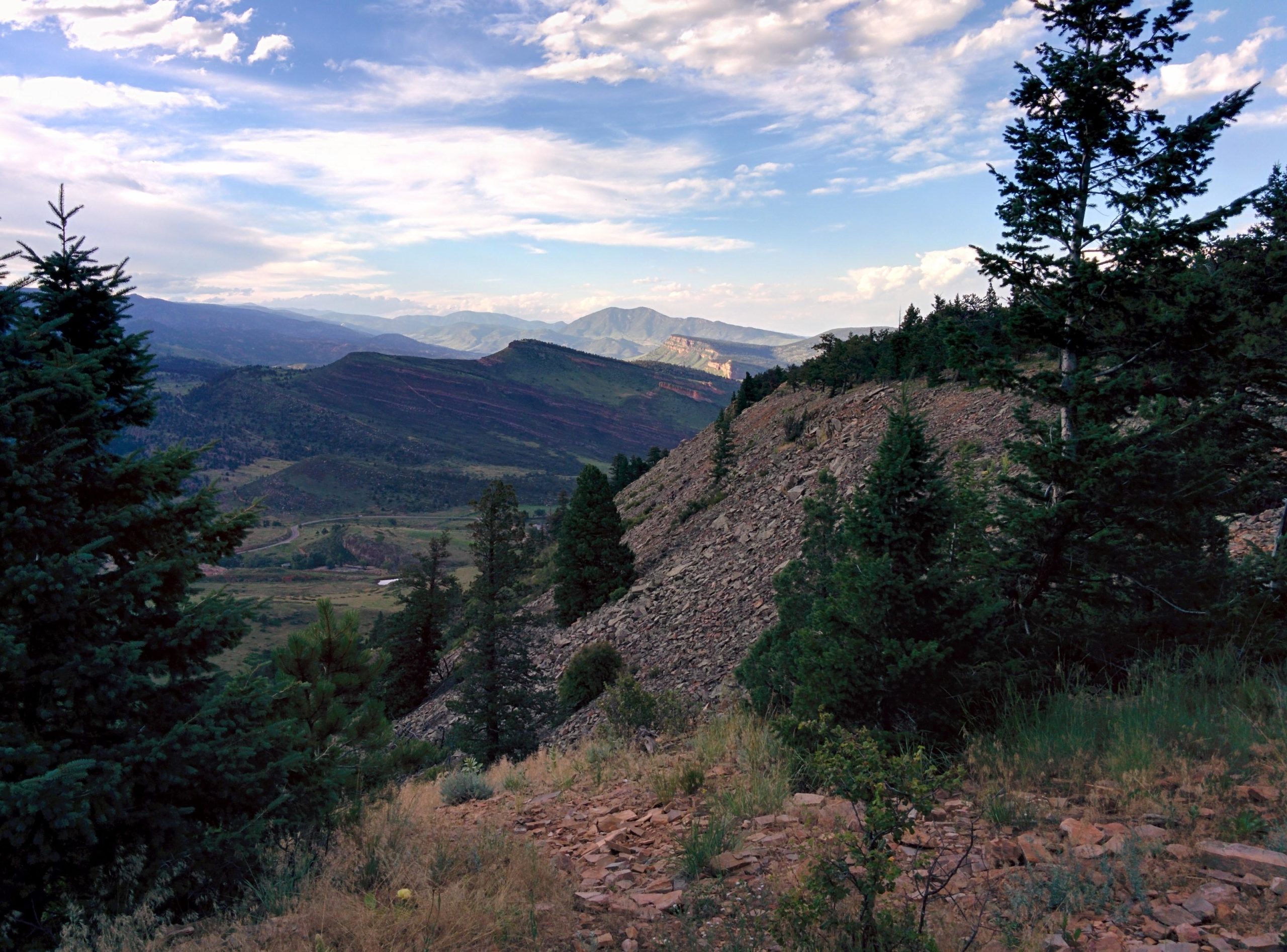 A scenic view of mountains under a partly cloudy sky, featuring rolling hills and a foreground of trees and rocky terrain, with a winding path visible in the lush valley below. Picture Rock Trail mountain bike trail.