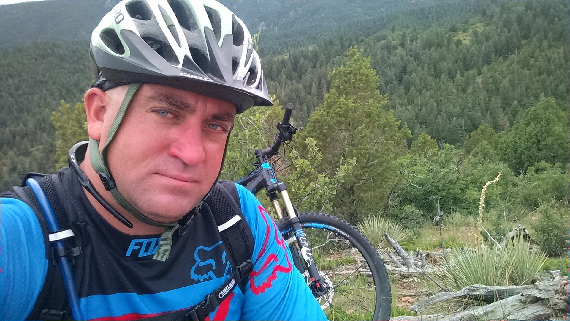 A man wearing a cycling helmet and a blue and black jersey stands near a mountain bike in a scenic outdoor setting, surrounded by trees and mountains in the background. Squirrel Creek mountain bike trail.