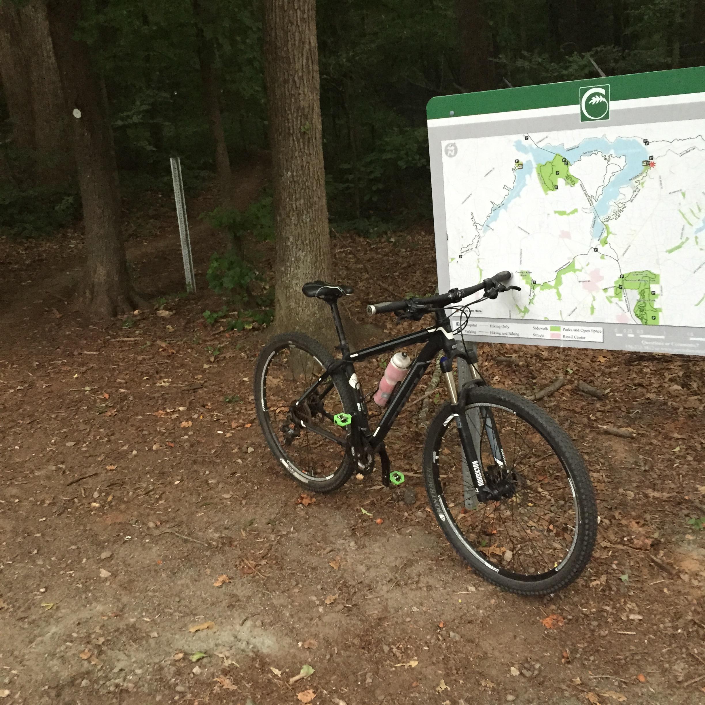 A mountain bike parked on a dirt path next to a trail map in a wooded area. The bike has a pink water bottle attached and is positioned near a sign displaying various trails and parks. The surrounding environment is lush with green trees and fallen leaves. Wild Turkey mountain bike trail.