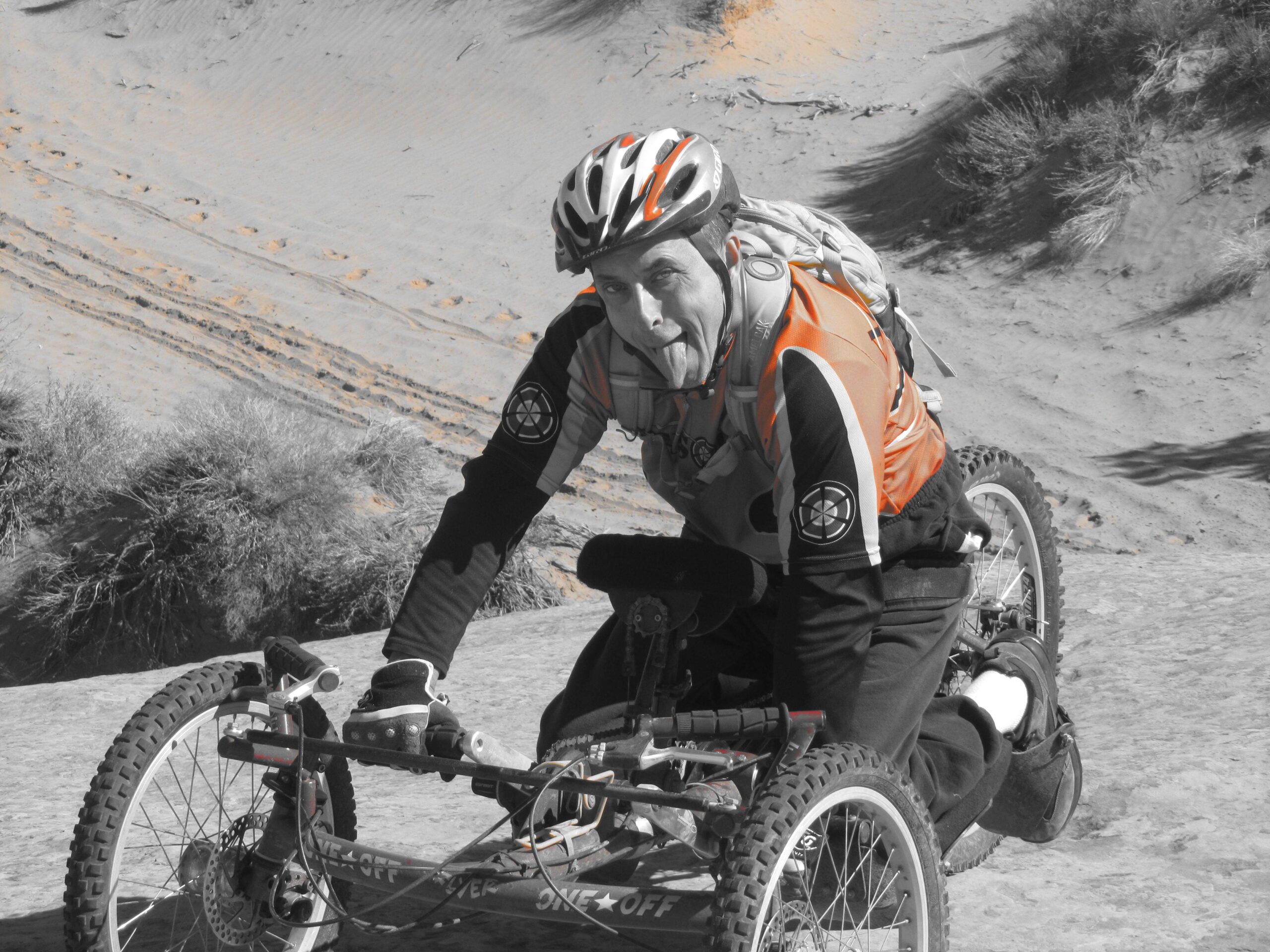 A person wearing a helmet and an orange cycling jersey is riding a specialized all-terrain cycle on a sandy surface. The background features gentle hills and scattered grass, with tracks visible in the sand. The image is primarily in black and white, highlighting the cyclist’s gear. Moab Brand Trails mountain bike trail.