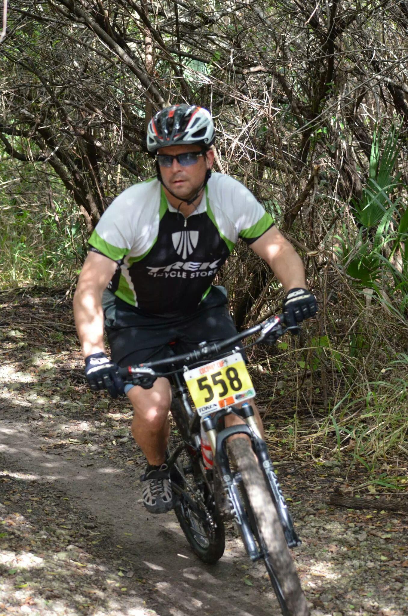 A mountain biker in a black and green jersey rides a bike along a dirt trail surrounded by dense vegetation. The biker wears a helmet and sunglasses, with a race number "558" visible on the front of their bike. Sunlight filters through the trees, highlighting the trail's natural surroundings. Caloosahatchee Regional Park mountain bike trail.