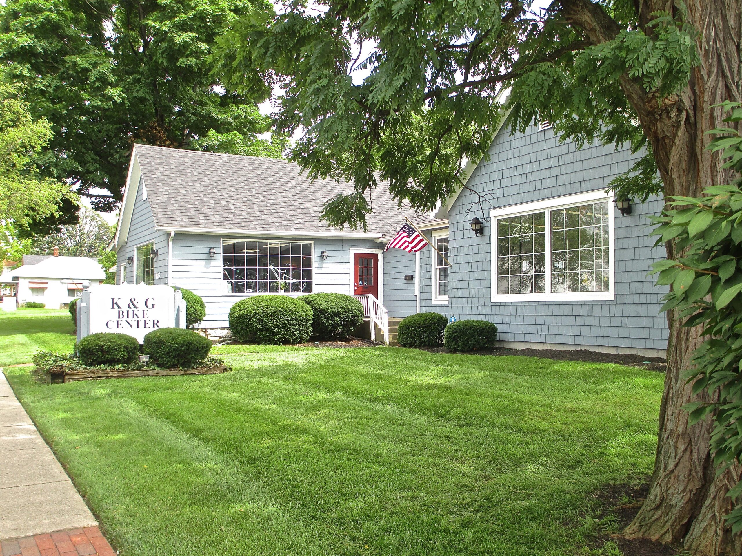 A light blue building with a shingled roof, featuring a red door and large windows. The front yard is well-maintained with green grass and round bushes. In front of the building, there is a sign that reads "K & G Bike Center." An American flag is displayed next to the entrance.