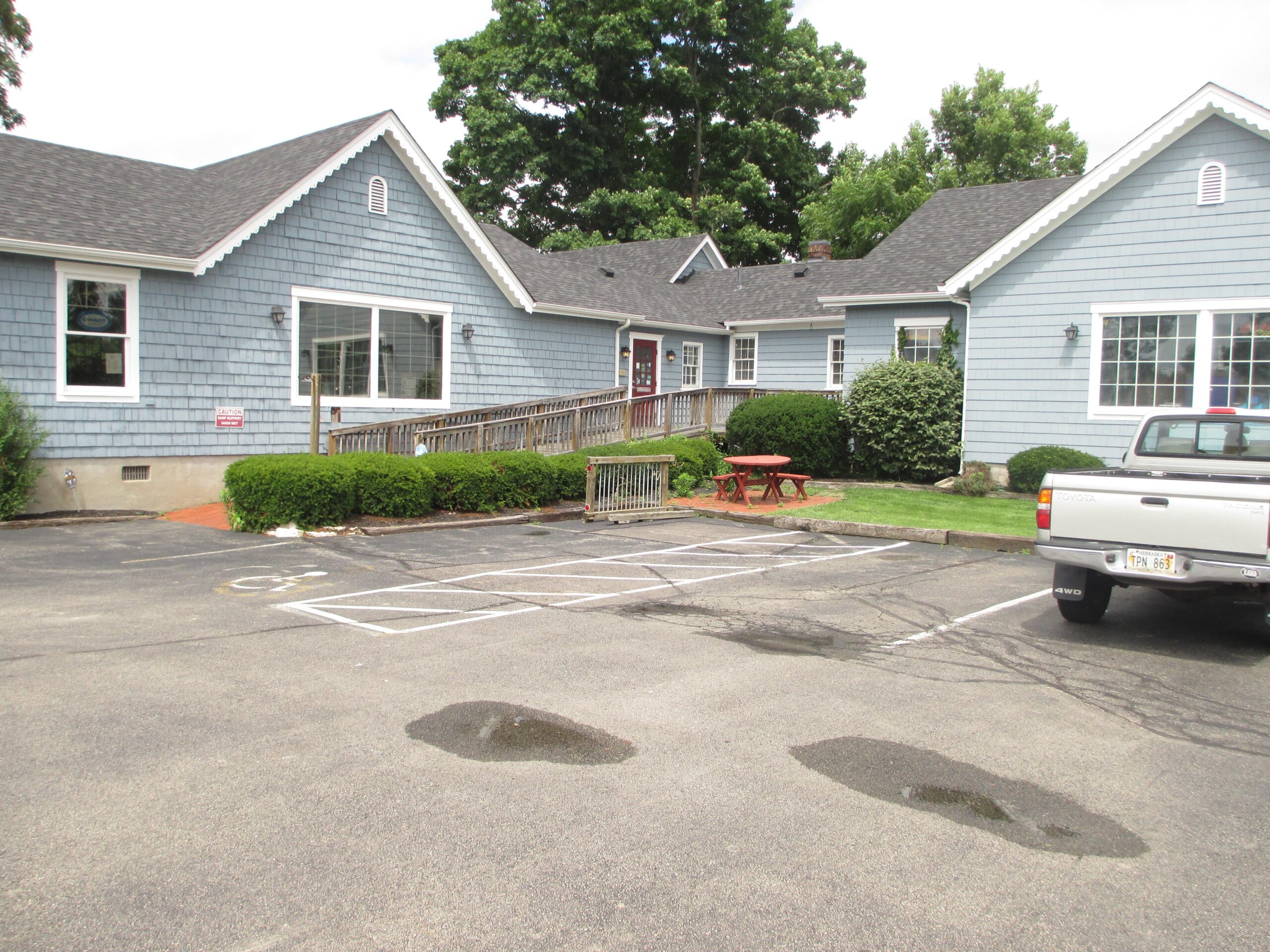 A light blue building with a shingle exterior, featuring a sloped roof and multiple windows. There is a wooden ramp leading to the entrance, surrounded by neatly trimmed bushes. In the foreground, a gravel parking lot is visible with several empty parking spaces. A picnic table can be seen on the lawn to the left, and a white pickup truck is parked nearby under a cloudy sky.