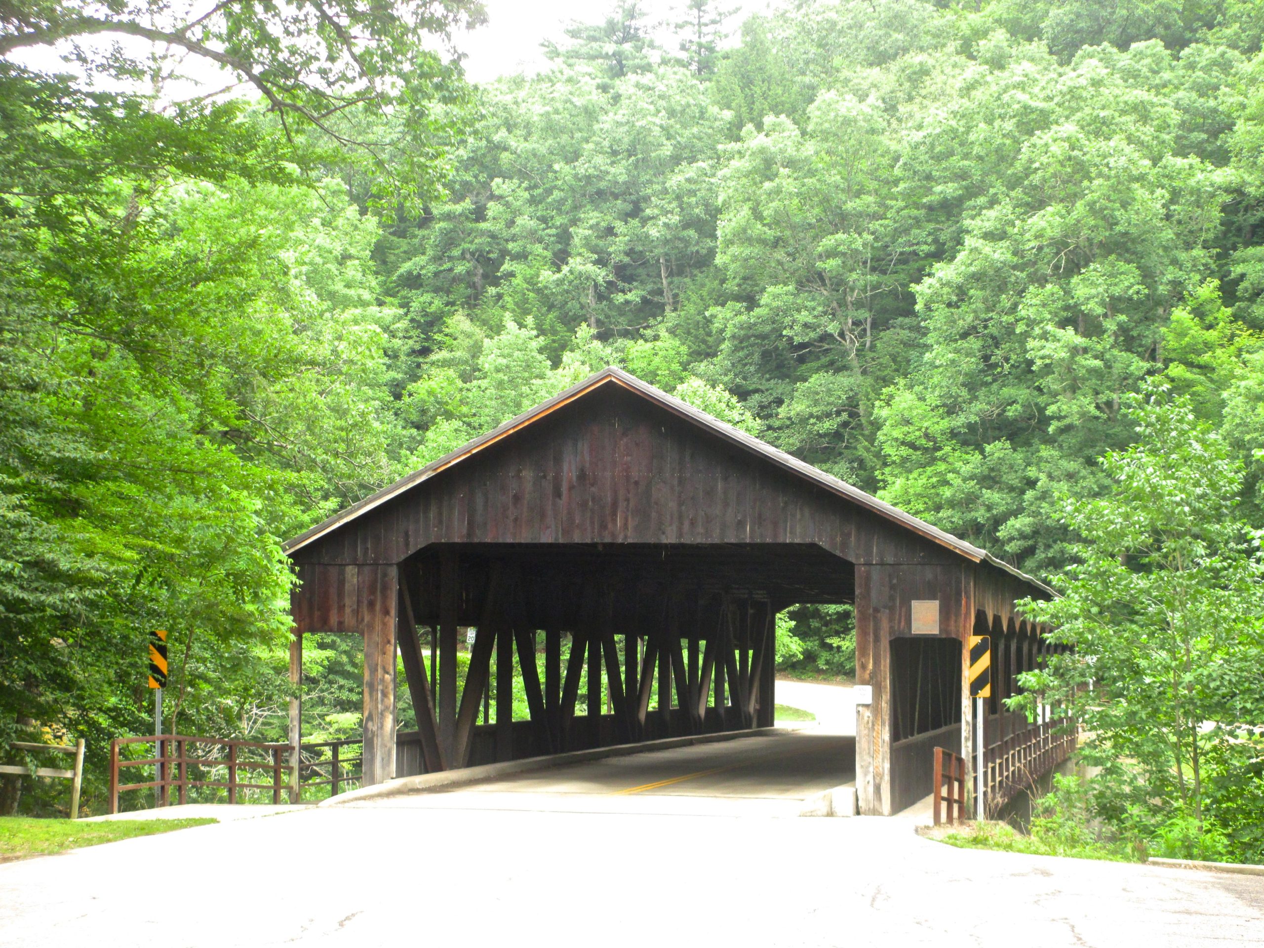 A rustic wooden covered bridge situated in a lush green landscape, surrounded by dense trees. The bridge spans a road that curves gently to the left, with a yellow diagonal warning sign visible near the entrance. Natural light filters through the foliage, creating a serene atmosphere. Mohican mountain bike trail.