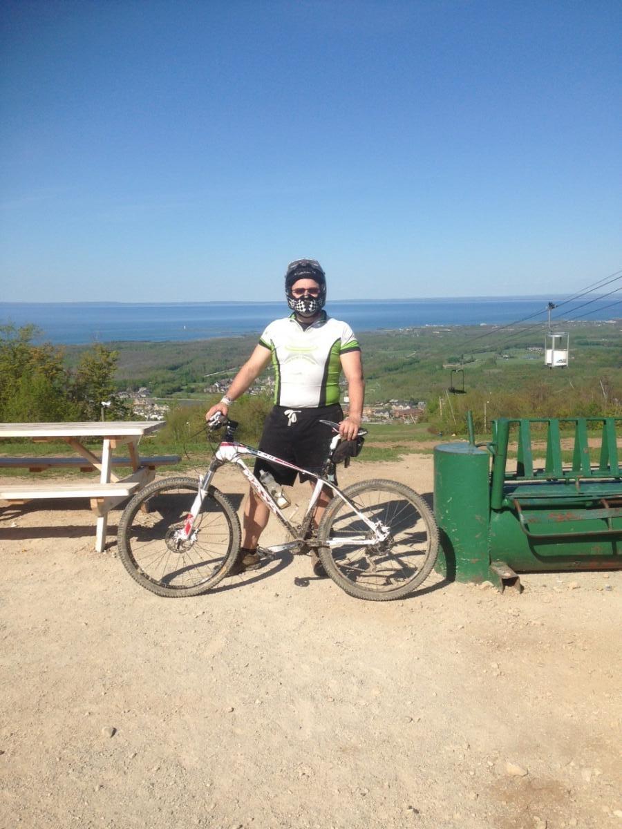 A mountain biker wearing a helmet and a cycling jersey stands next to a white mountain bike on a dirt trail. In the background, there is a panoramic view of a lake and green hills under a clear blue sky. A bench and a ski lift can also be seen in the area.