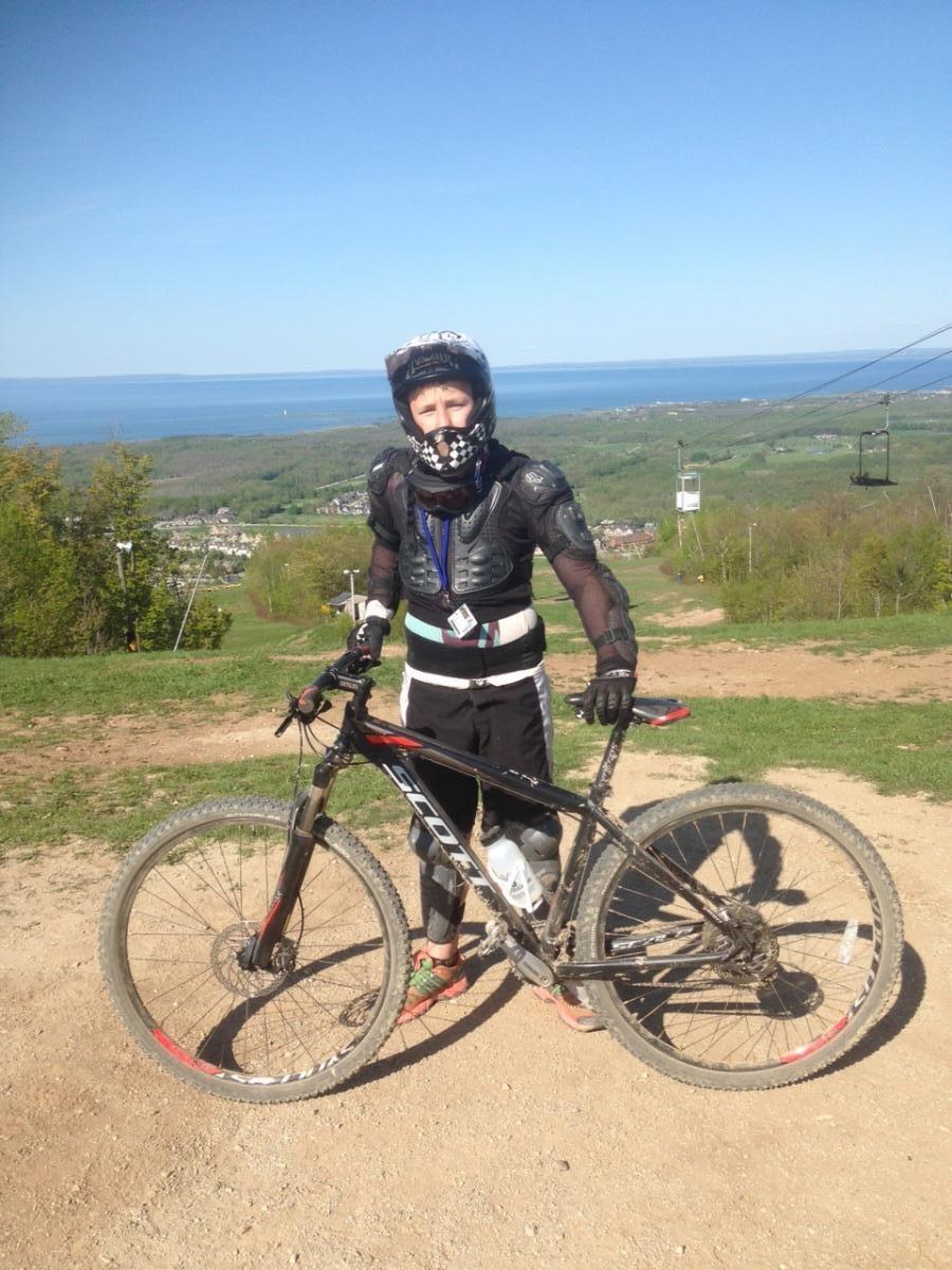 A young mountain biker wearing protective gear, including a helmet and knee pads, stands beside a black mountain bike. The background features a scenic view of rolling green hills and a large body of water under a clear blue sky. The biker has a focused expression and is positioned on a dirt path, suggesting a mountain biking trail.