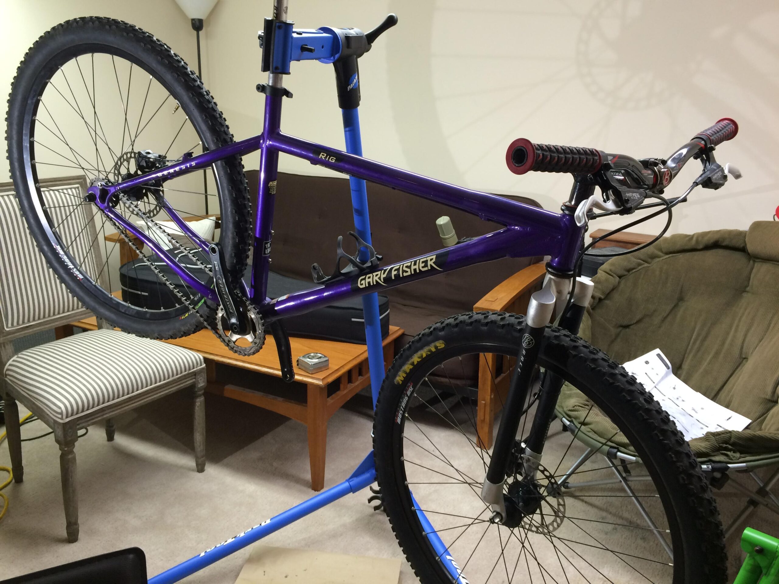 Gary Fisher Rig 29er: A purple Gary Fisher mountain bike frame is displayed on a blue bike stand, with its front wheel elevated. The bike features black tires and grips, and there's a striped chair and a round green chair in the background, along with a wooden table and some papers.