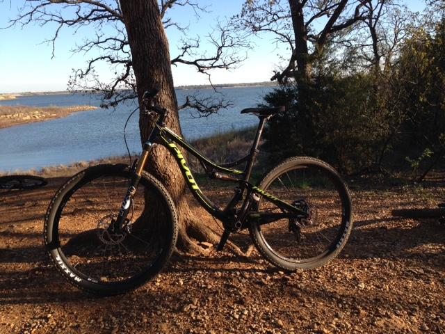 Pivot Mach 4: A mountain bike resting against a tree near a lake, surrounded by rocky terrain and sparse greenery under a clear blue sky.