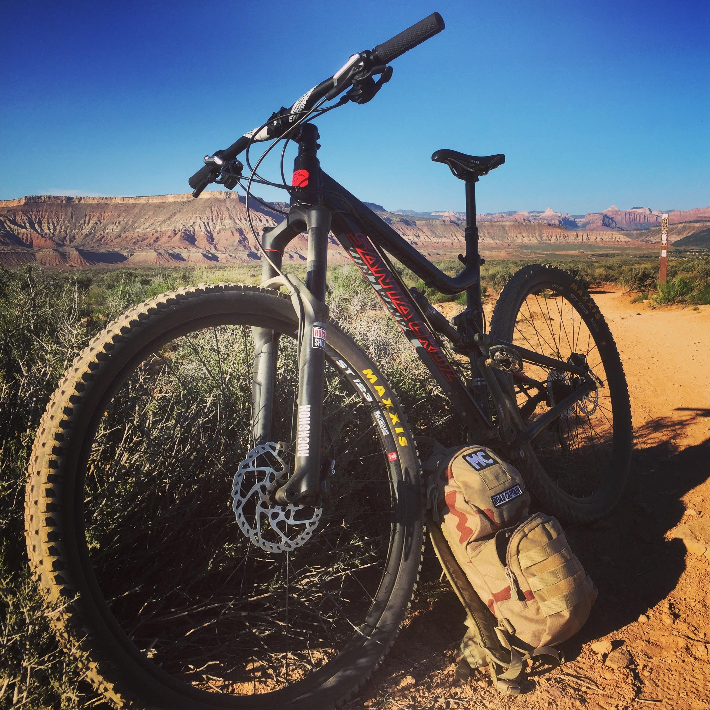Santa Cruz Tallboy LT: A mountain bike parked alongside a sandy trail, set against a backdrop of rocky cliffs and clear blue skies. A small, camouflaged backpack rests on the ground next to the bike. The scene captures the essence of outdoor adventure and biking exploration.