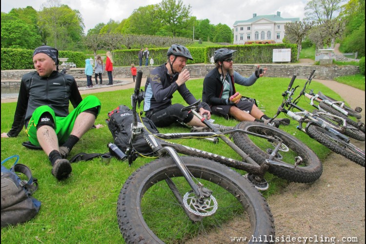 Three mountain bikers resting on the grass after a ride, with two bicycles lying nearby. One cyclist in green shorts is sitting and appearing relaxed, while another is enjoying a snack. The third cyclist is holding a phone, possibly taking a photo. In the background, a group of people can be seen near a fountain and a large building surrounded by greenery.