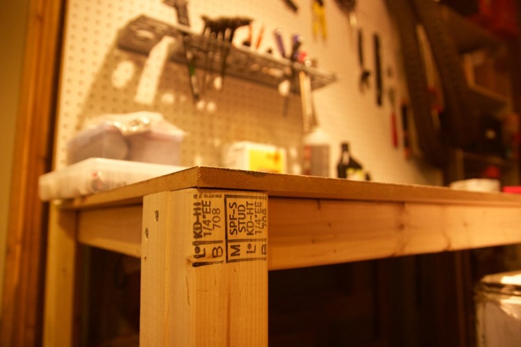A close-up view of a wooden workbench corner with visible markings on the wood, set against a background of tools and storage containers on a pegboard wall. The image captures the texture of the wood and the organized workspace atmosphere.