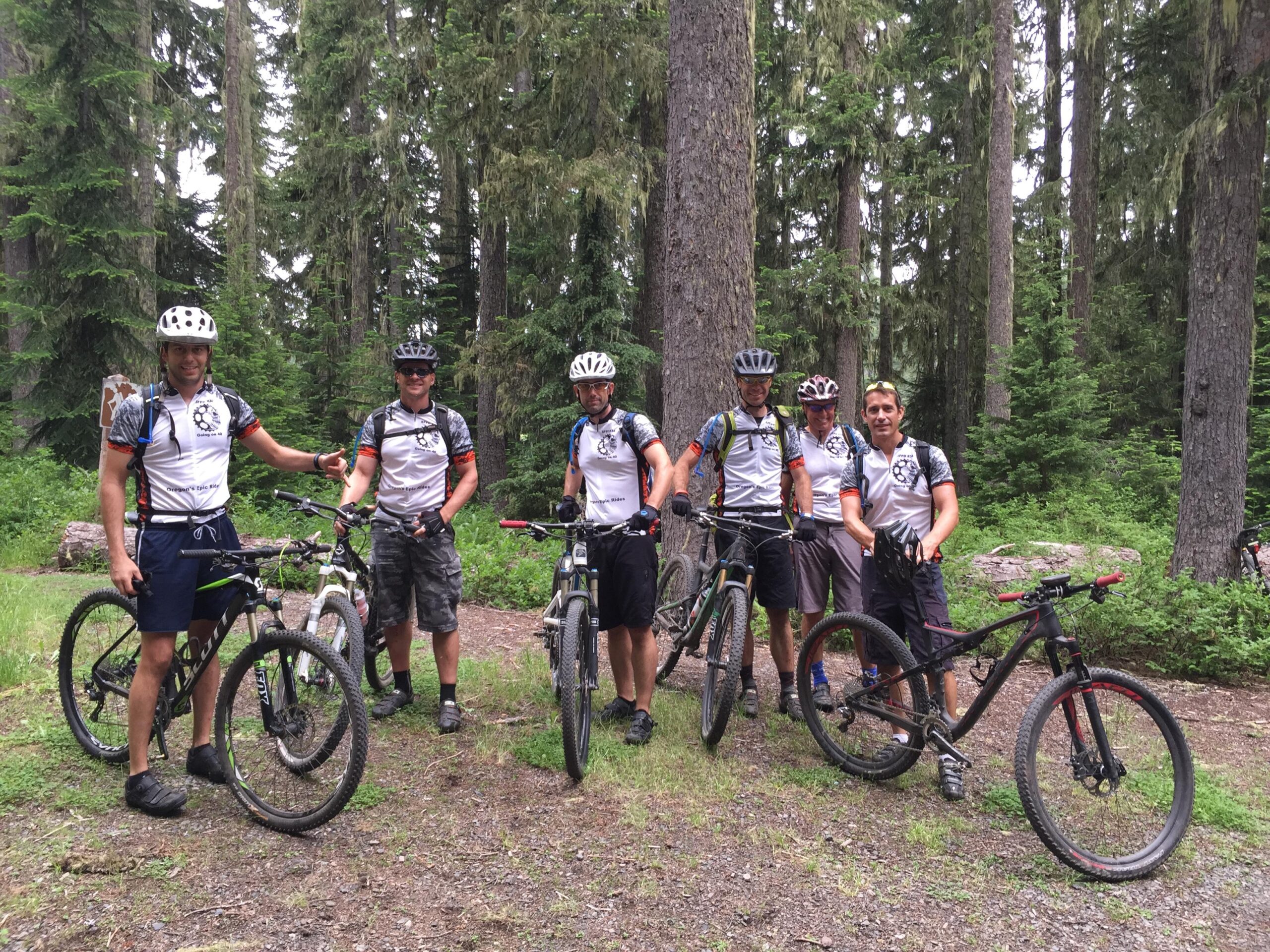 A group of six mountain bikers stands in a forested area, smiling and posing with their bikes. They are wearing matching jerseys and helmets, surrounded by tall trees and greenery. The atmosphere is casual and friendly, suggesting a day of biking in nature. Middle Fork Trail mountain bike trail.