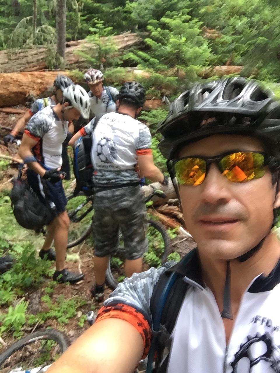 A group of mountain bikers pause in a dense forest setting, surrounded by lush greenery and fallen logs. One biker is taking a selfie while the others gather around their bikes, dressed in cycling gear and helmets. Middle Fork Trail mountain bike trail.