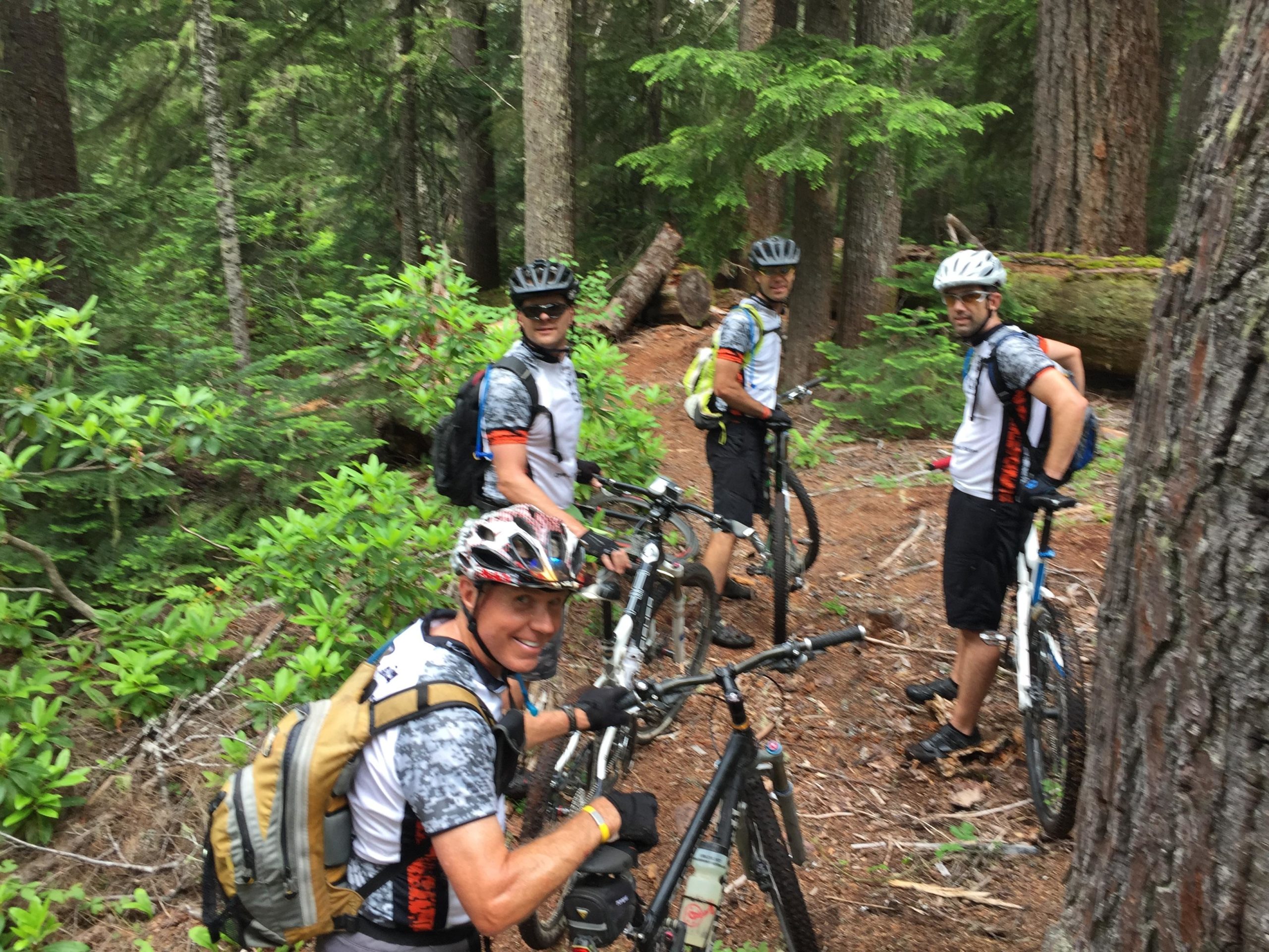 Four mountain bikers pause on a wooded trail, surrounded by tall trees and lush greenery. They are wearing matching jerseys and helmets, looking towards the camera with smiles. Their bikes are parked nearby, and the scene captures the spirit of outdoor adventure and camaraderie in nature. Middle Fork Trail mountain bike trail.