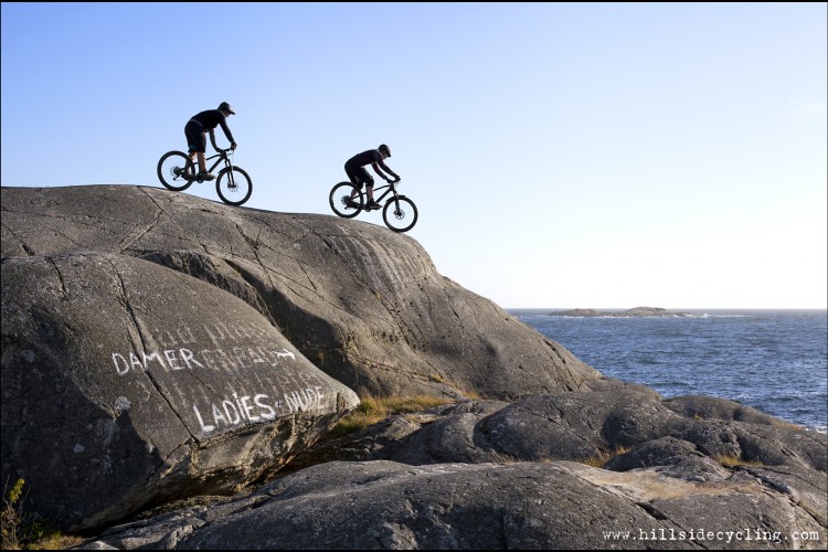 Two mountain bikers riding on a rocky outcrop overlooking the sea. A white painted sign on the rock indicates a trail for ladies, with arrows pointing in the direction of the path. The scene is set against a clear sky and the ocean in the background.