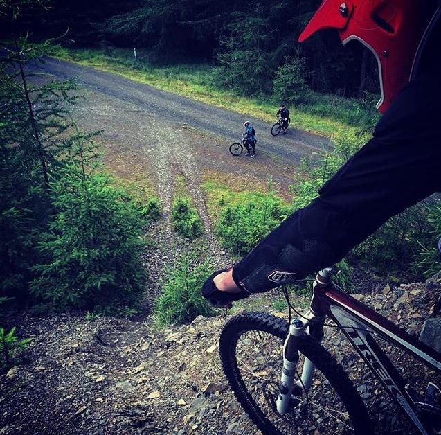 A mountain biker in protective gear looks down a gravel path in a forested area, with two other bikers in the background. The scene captures the excitement and anticipation of downhill cycling amidst lush greenery. Innerleithen mountain bike trail.