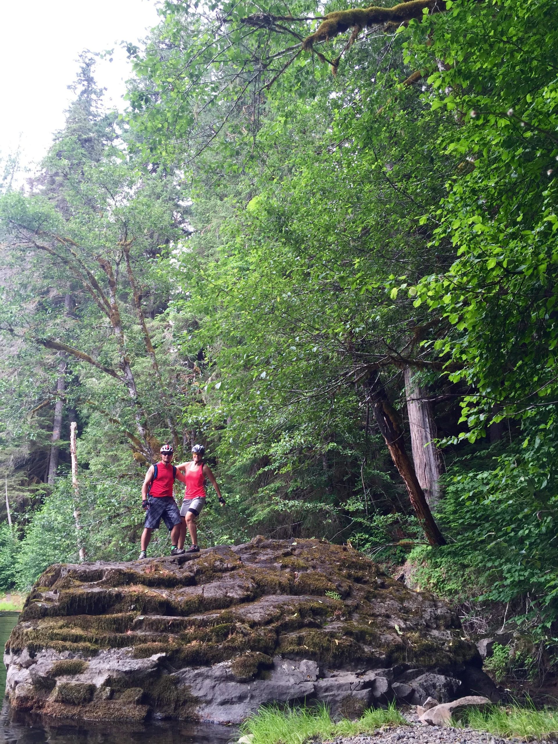 Two individuals standing on a large rock in a lush green forest. They are wearing red shirts and biking gear, with trees and foliage surrounding them. A calm body of water is visible near the rock, adding to the natural setting. Lewis River mountain bike trail.