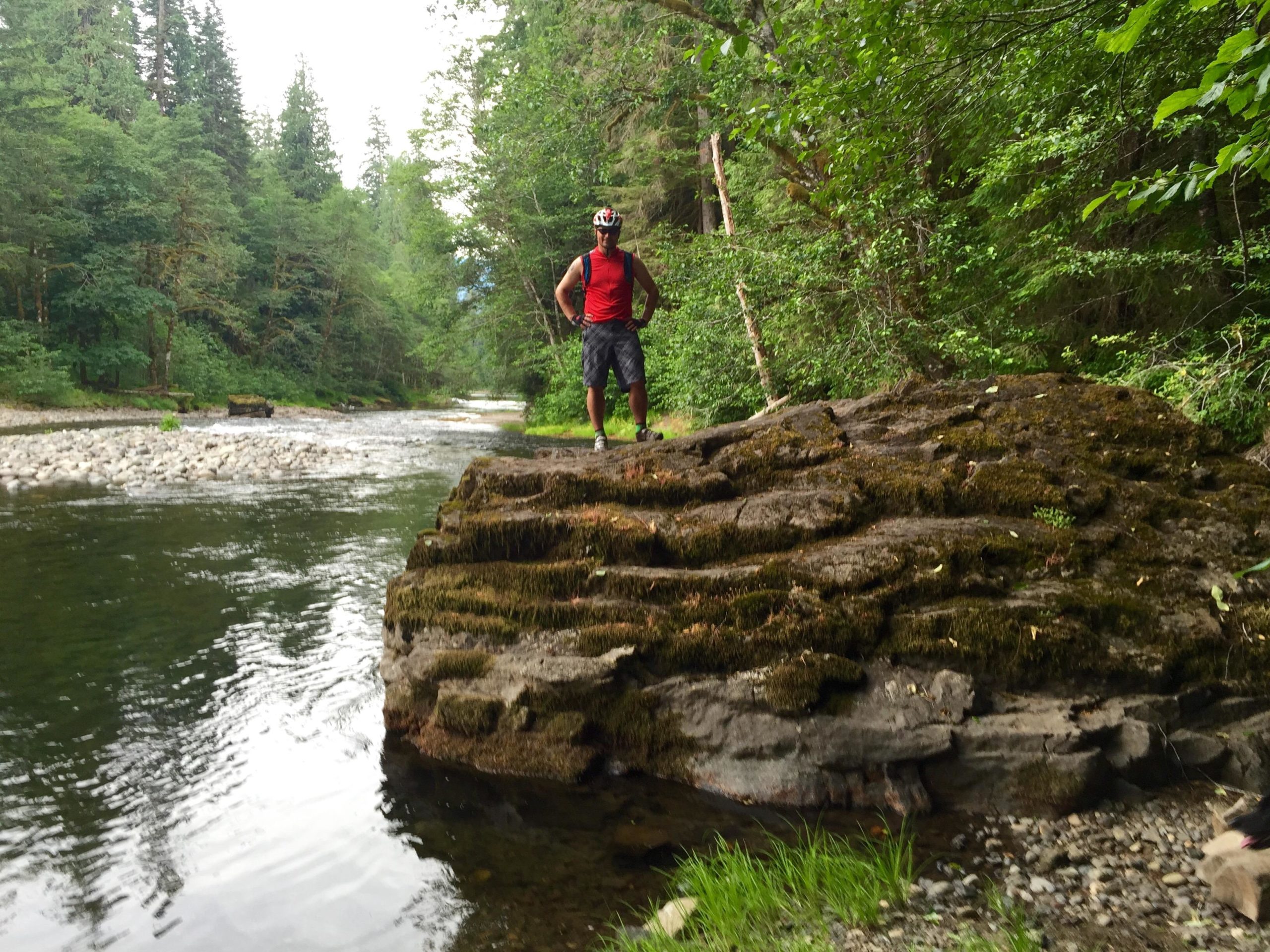 A person in a red shirt stands on a moss-covered rock by a flowing river, surrounded by lush green trees and a natural landscape. The water reflects the greenery, and smooth pebbles line the riverbank. The scene conveys a sense of tranquility and connection to nature. Lewis River mountain bike trail.