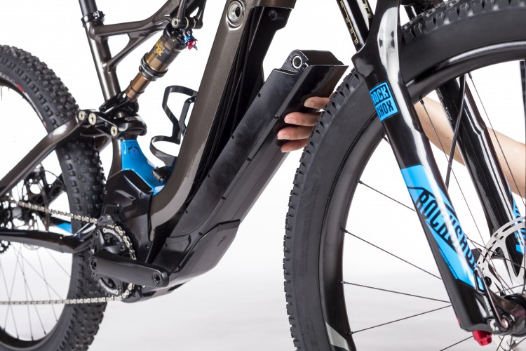 Close-up view of an electric mountain bike showing the motor and battery compartment, with a hand reaching to adjust or remove the battery. The bike features a black frame, blue accents, and a RockShox front fork. The background is plain white to enhance focus on the bike's components.