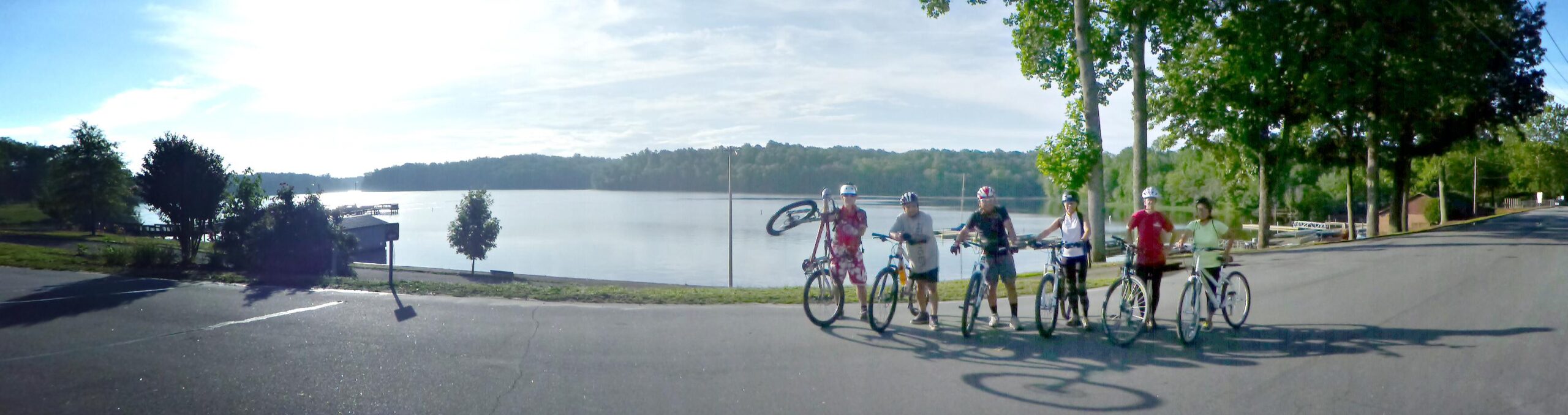 A group of five cyclists posing on a paved road by a lake, with trees and greenery in the background. The sun is shining, and the water reflects the light. Some cyclists hold their bikes while others have their bikes next to them, showcasing a casual and friendly atmosphere. Salem Lake mountain bike trail.