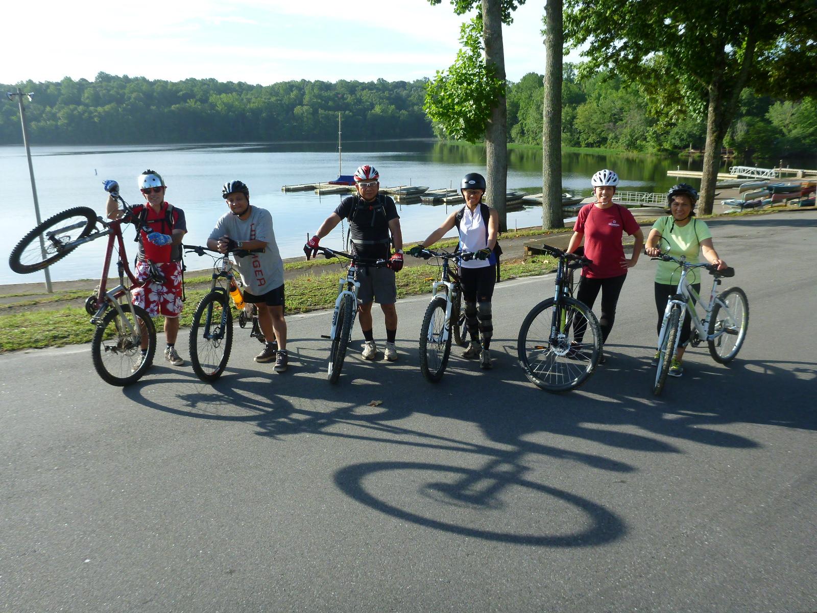 A group of six cyclists posing with their mountain bikes near a serene lake. They are wearing helmets and a mix of casual cycling attire. A few bikes are held aloft, while the backdrop features lush greenery and a calm water surface. The scene suggests a day of outdoor activity and camaraderie. Salem Lake mountain bike trail.