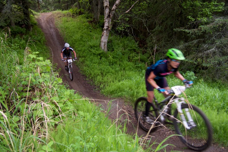 Two mountain bikers are navigating a winding dirt trail surrounded by lush greenery. One rider is seen in focus, with a bright green helmet, and appears to be moving quickly, while the other biker is slightly blurred in the background. The scene captures the excitement of mountain biking in a natural setting.