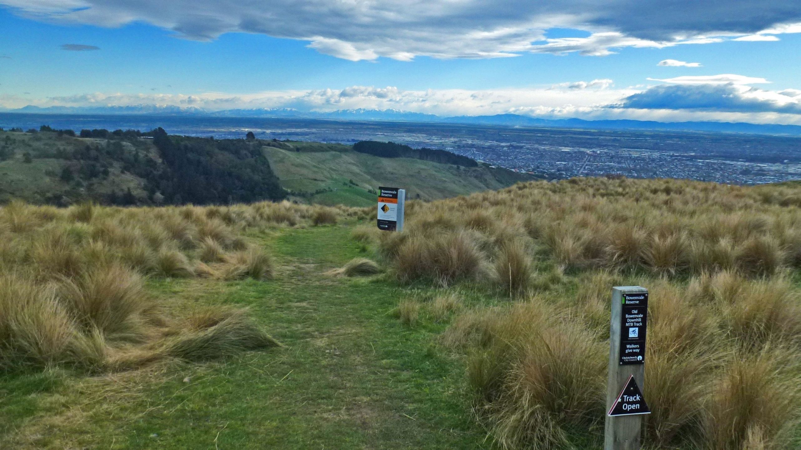 A scenic view from a hiking trail overlooking rolling hills and a distant cityscape, with signs indicating trail options and recommendations. The grassy terrain is dotted with tall grass, and the sky is partly cloudy, adding to the serene atmosphere of the natural setting. Bowenvale Downhill mountain bike trail.