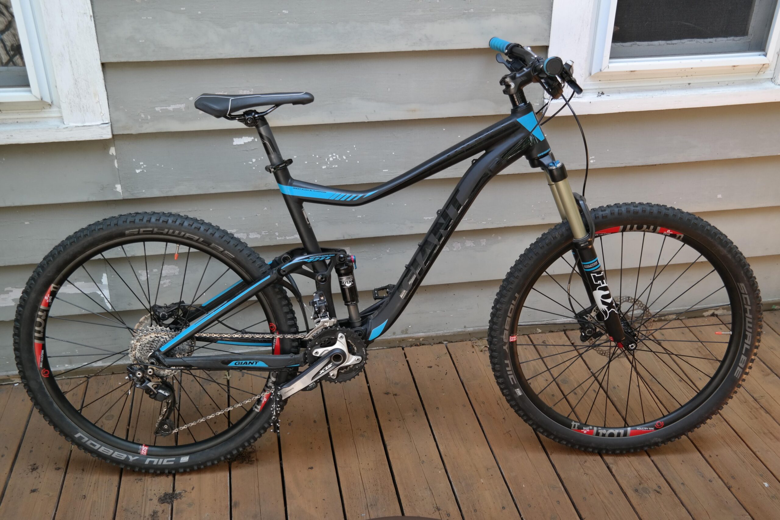 Giant Trance 27.5 2: A black mountain bike with blue accents is parked on a wooden deck. The bike features a sturdy frame, dual suspension, wide tires with knobby tread, and a front shock absorber. In the background are gray wooden siding and a window, hinting at a home setting.