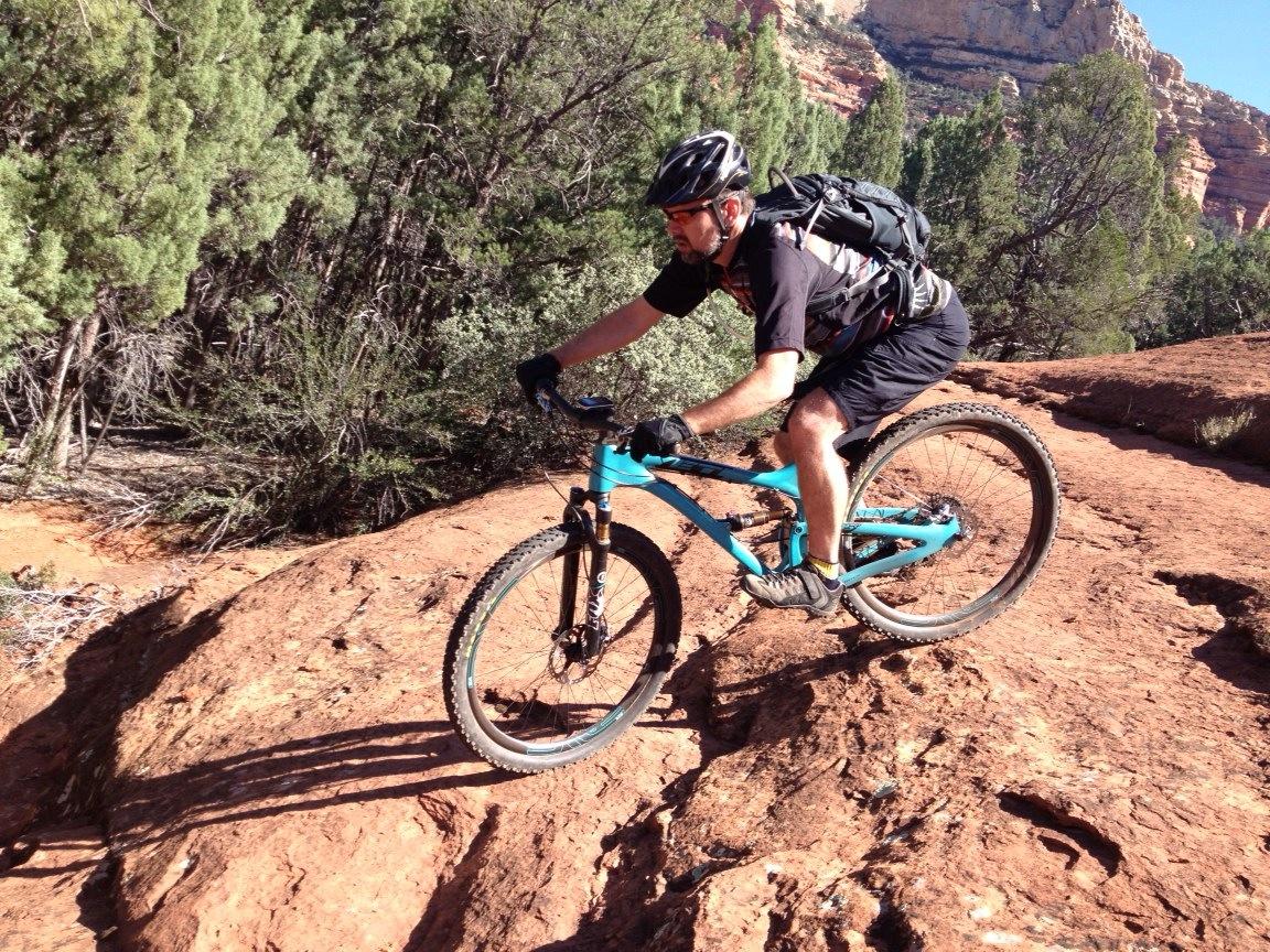 Yeti SB95: A mountain biker navigating rocky terrain in a forested area, wearing a helmet and backpack. The sun casts shadows on the textured red rocks, surrounded by greenery.