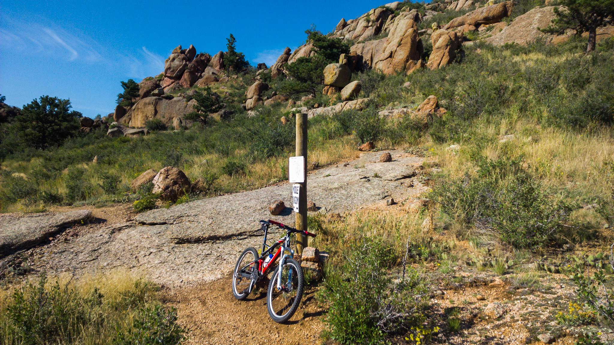 A mountain bike rests against a wooden signpost along a rocky trail, surrounded by tall grass and sparse vegetation. In the background, rugged rock formations rise against a clear blue sky. The scene captures a tranquil outdoor setting ideal for biking and hiking. Curt Gowdy State Park mountain bike trail.