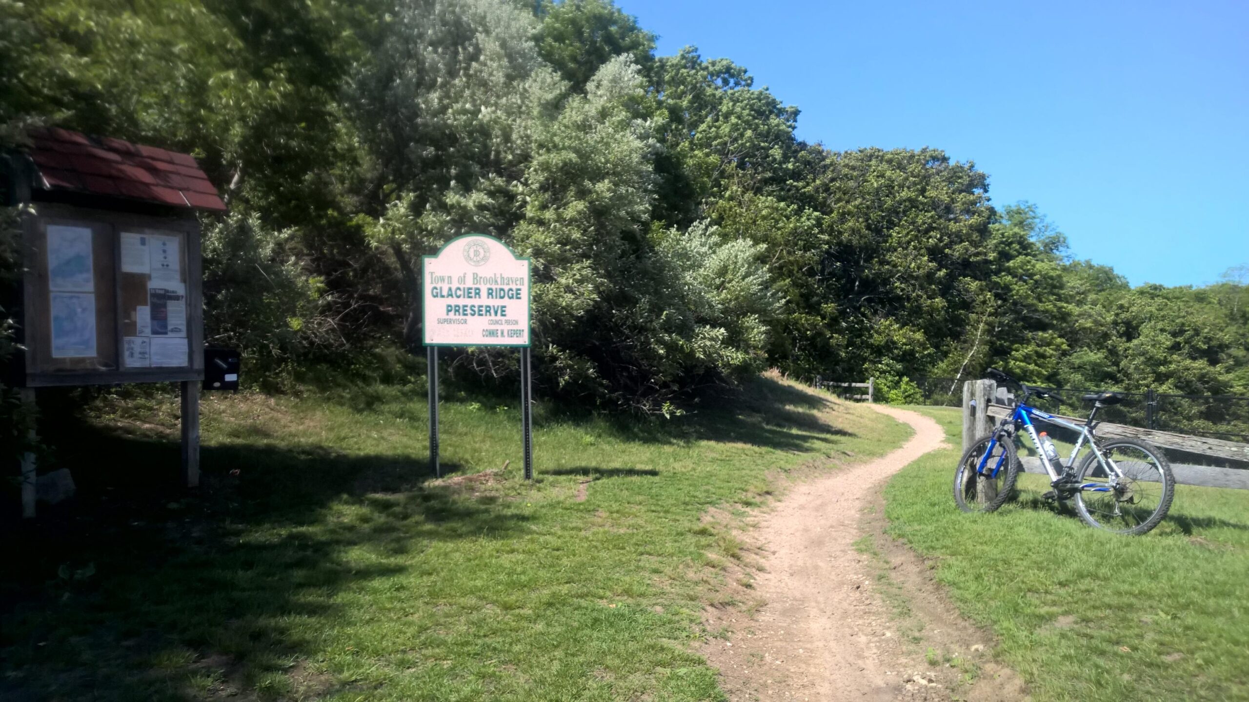 Trek 4300: A sign marking the entrance to Glacier Ridge Preserve, surrounded by lush greenery. A dirt path leads from the sign, and a blue bicycle is parked nearby on the grass. The sky is clear and sunny.