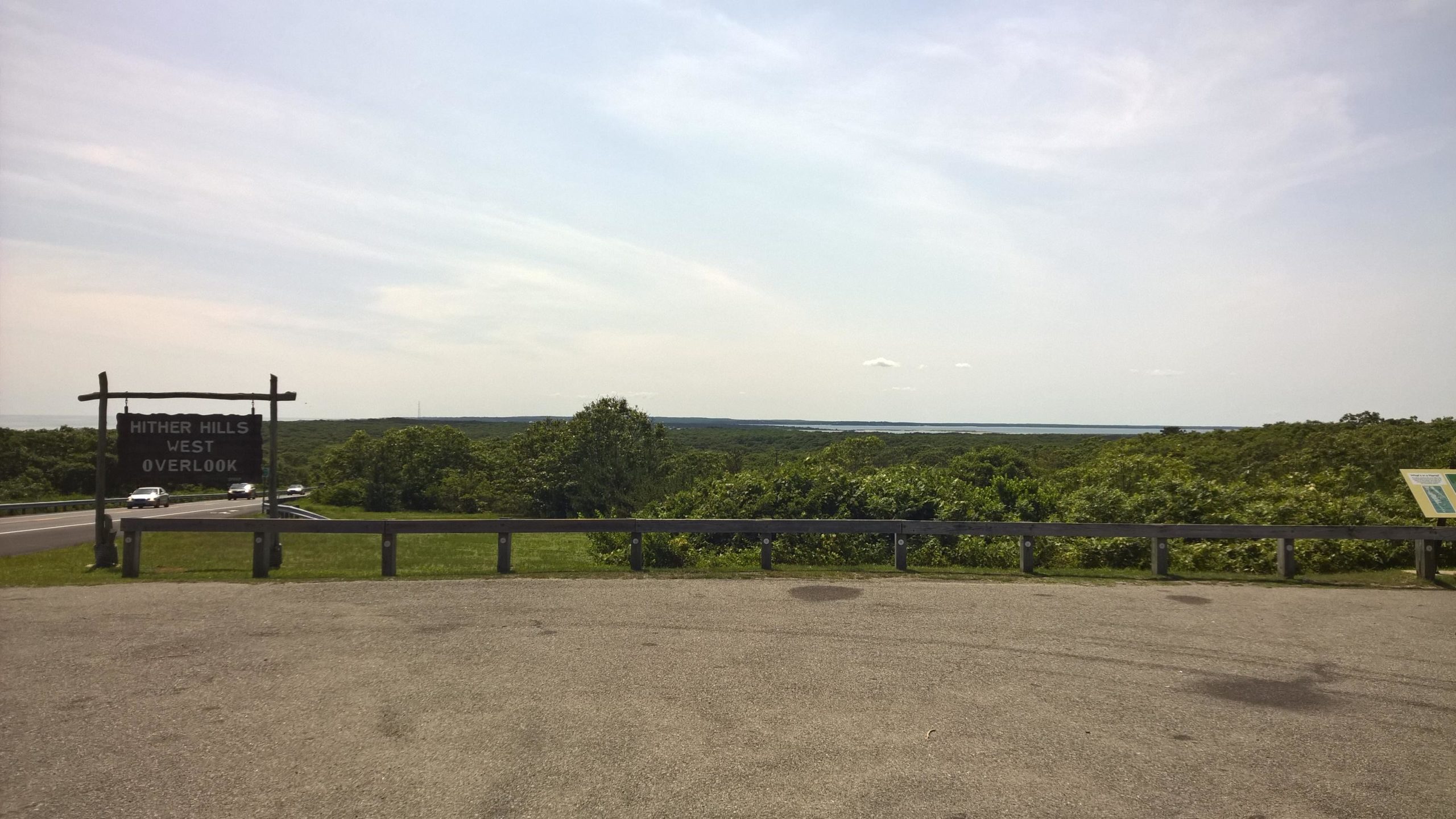 Sign for Hither Hills West Overlook in a scenic area with lush greenery and distant water views. A paved area is in the foreground, and vehicles can be seen on the road nearby under a clear sky. Hither Woods mountain bike trail.