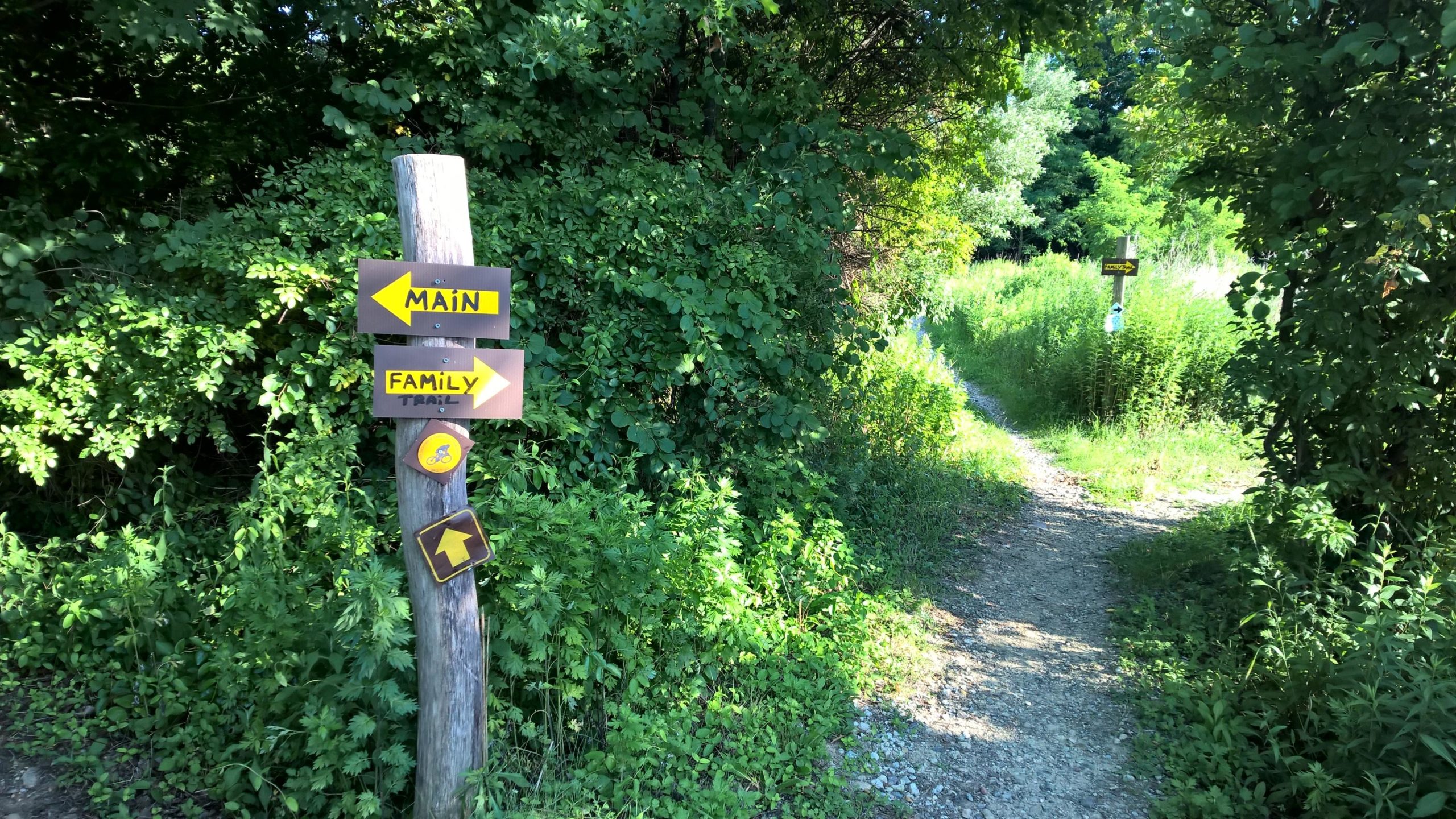 Wooden post with directional signs indicating "MAIN" and "FAMILY TRAIL," surrounded by lush green vegetation. A gravel path leads into the greenery, providing access to the trails. Rocky Point mountain bike trail.