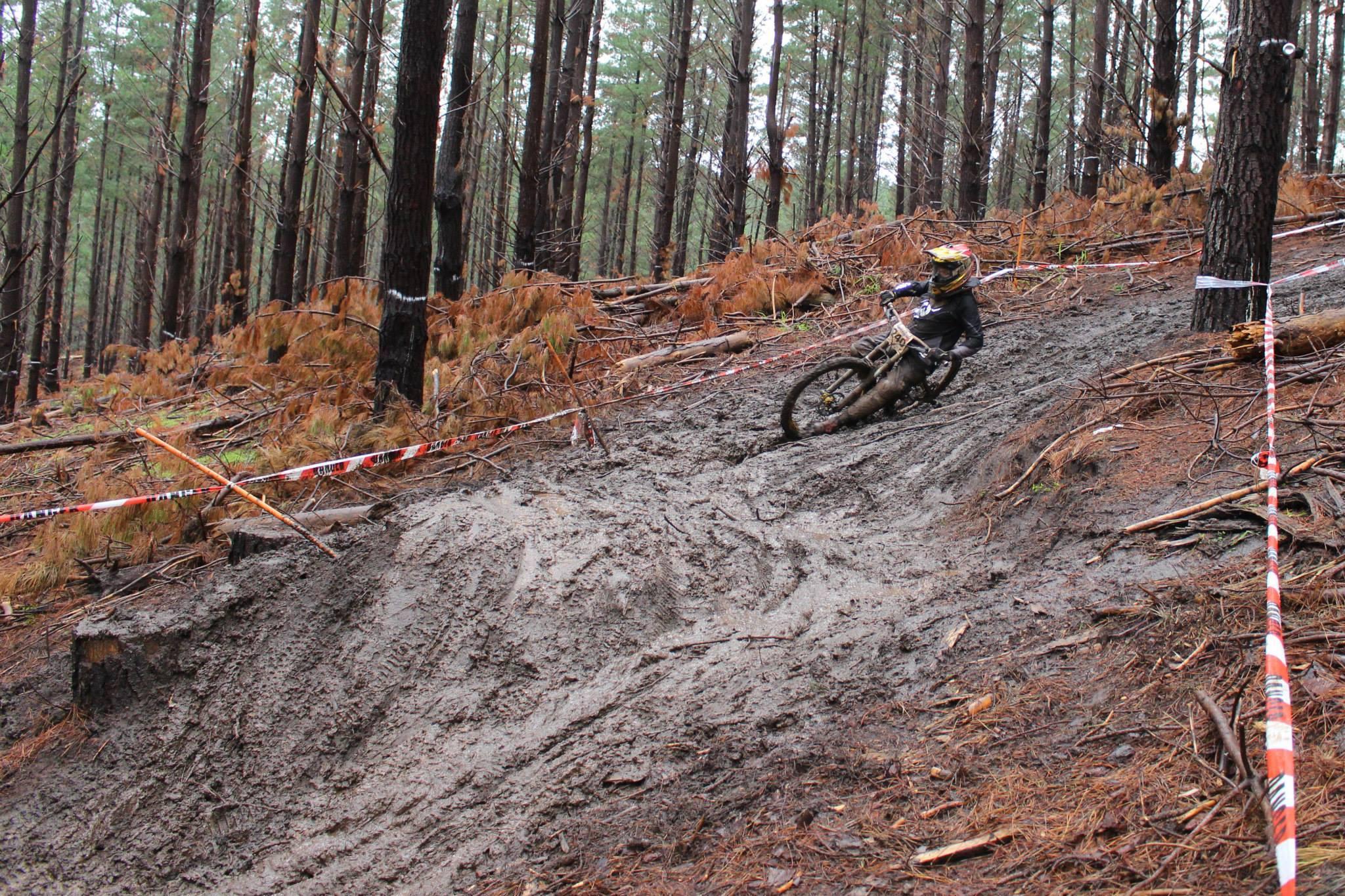 A mountain biker navigating a muddy trail in a dense forest, with tall trees in the background and orange and white caution tape marking the path. The rider is leaning into a turn, showcasing skill and balance on a wet, slippery surface. Wellington Mills mountain bike trail.