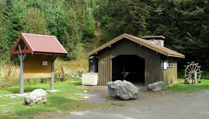 A wooden shelter with a dark roof and an open front is situated in a grassy area, surrounded by trees. Nearby is a signboard with a red roof and a blank space for postings. A large rock is positioned in front of the shelter, alongside a water wheel structure. A dirt path leads into the woods in the background. Craigieburn Forest mountain bike trail.