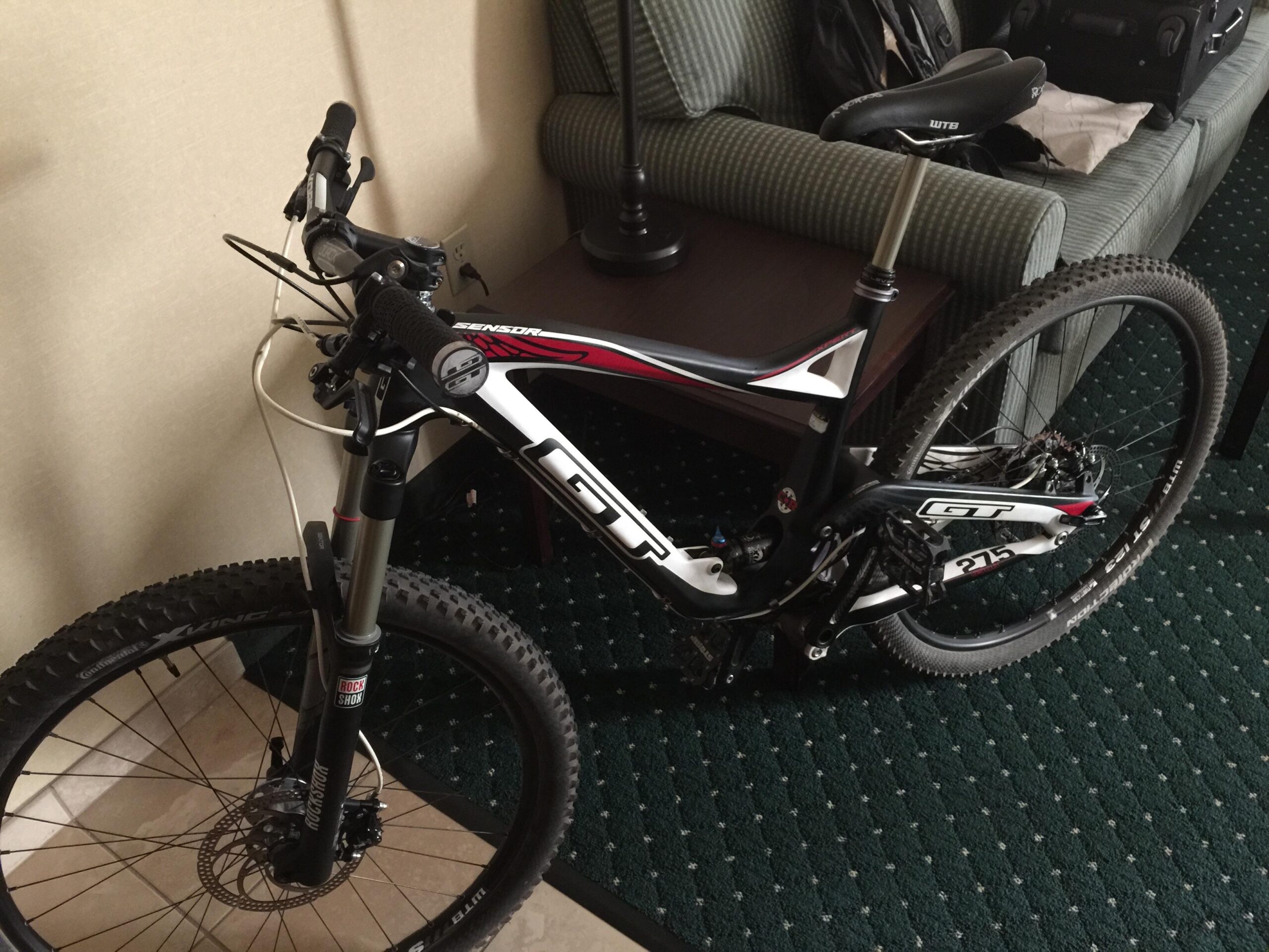 GT Sensor Carbon Expert: A mountain bike with a black and white frame is positioned in a room. The bike features thick tires, a front suspension fork, and a prominent logo. In the background, there's a green carpet, a couch, and a table with a lamp. Luggage and a backpack are also visible beside the couch.