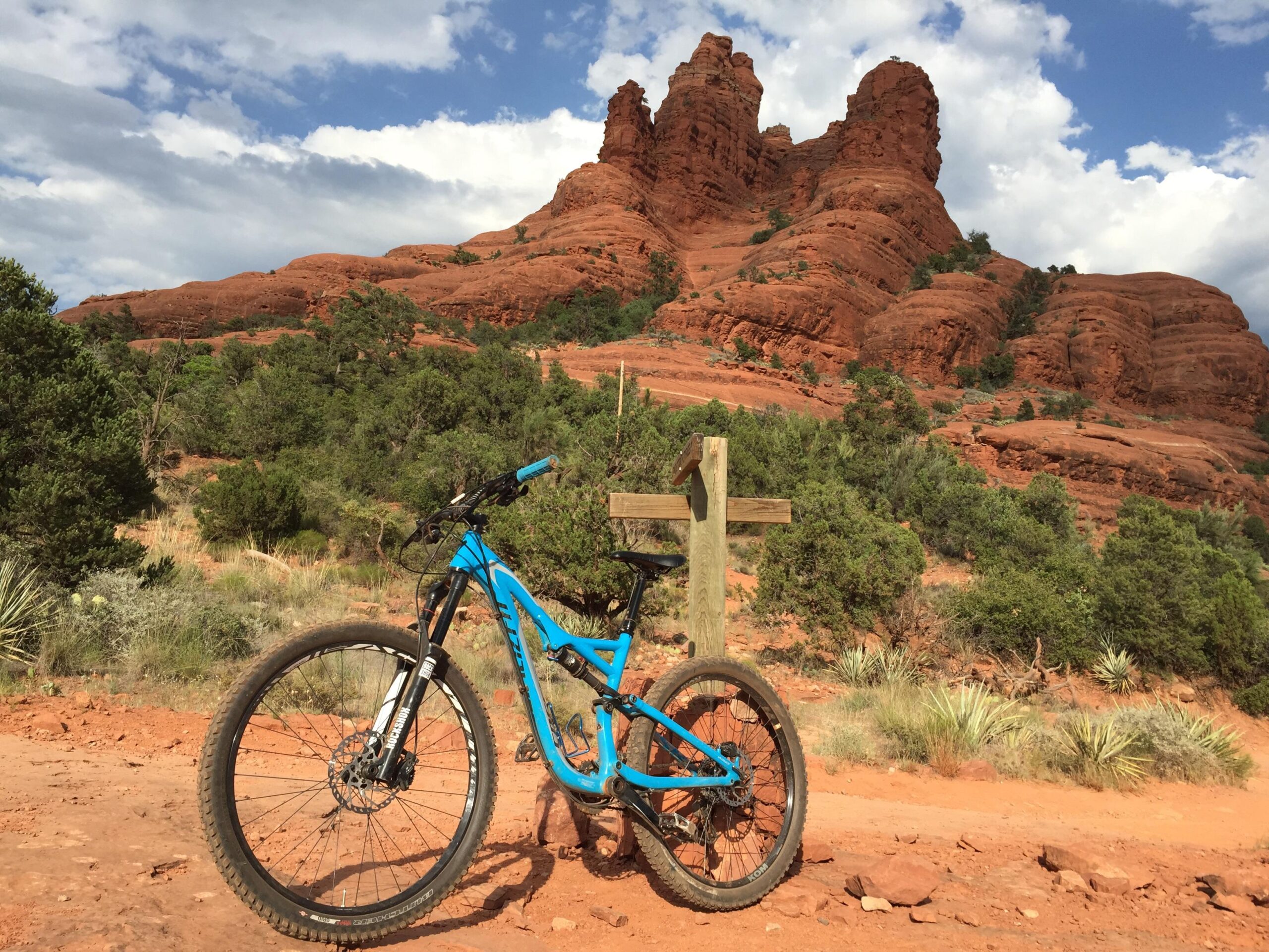 Specialized Stumpjumper FSR Comp EVO: A vibrant blue mountain bike stands on a dirt path in front of striking red rock formations under a partly cloudy sky. Lush greenery surrounds the area, highlighting the natural beauty of the landscape. A wooden signpost is visible next to the bike, enhancing the outdoor adventure theme of the scene.