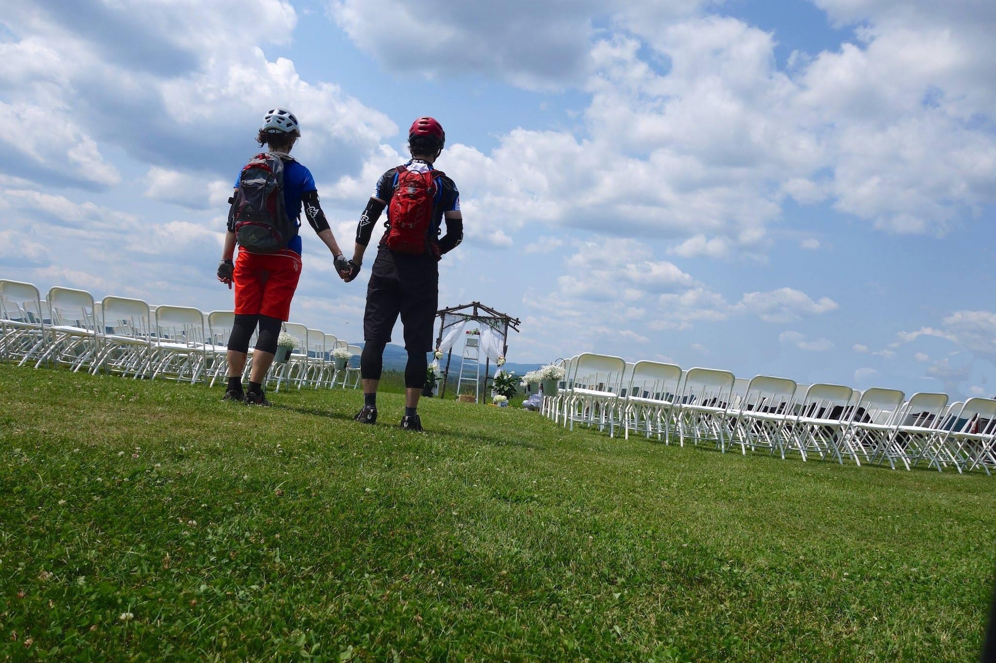 Two cyclists holding hands while standing on a grassy area, looking towards a wedding setup with rows of chairs and a decorated arch in the background. The sky is partly cloudy, suggesting a pleasant outdoor atmosphere. Kingdom Trails mountain bike trail.