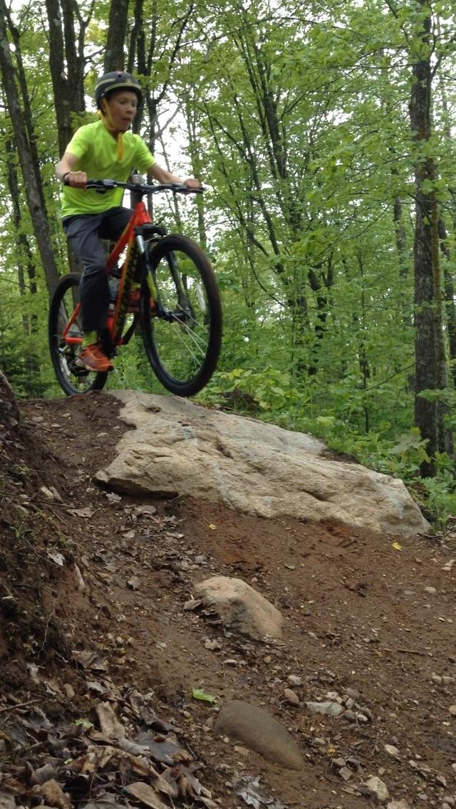 A young boy in a bright yellow shirt and helmet performs a bicycle jump off a large rock in a wooded area, surrounded by lush green trees. The bike's front wheel is off the ground, showcasing an action-packed moment in mountain biking. WinMan Trails mountain bike trail.