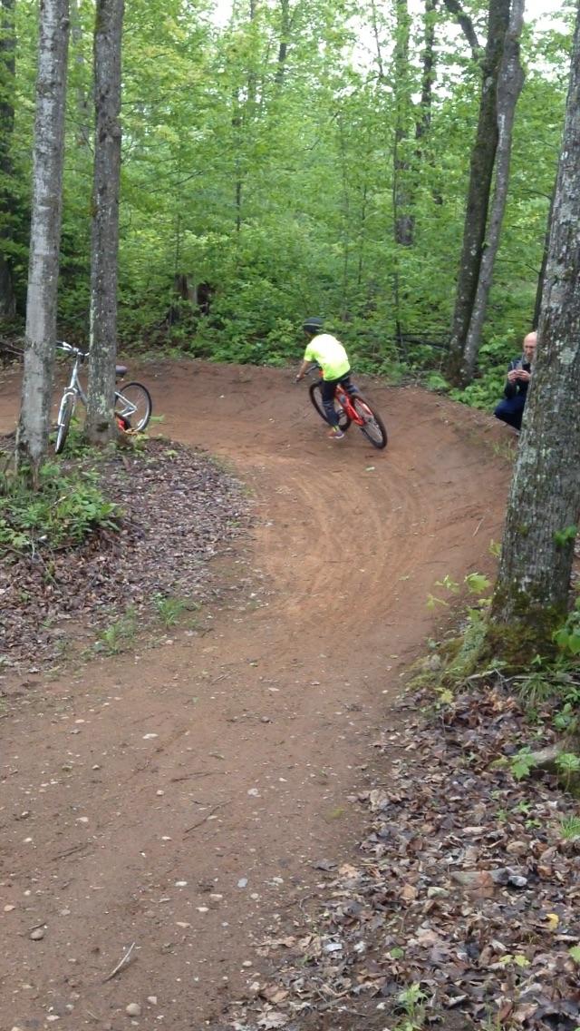 A child in a bright yellow shirt rides a bicycle around a dirt trail in a wooded area, with trees and greenery surrounding the path. Another bicycle is parked nearby, and an adult is seen taking a photo as the child leans into the turn. WinMan Trails mountain bike trail.