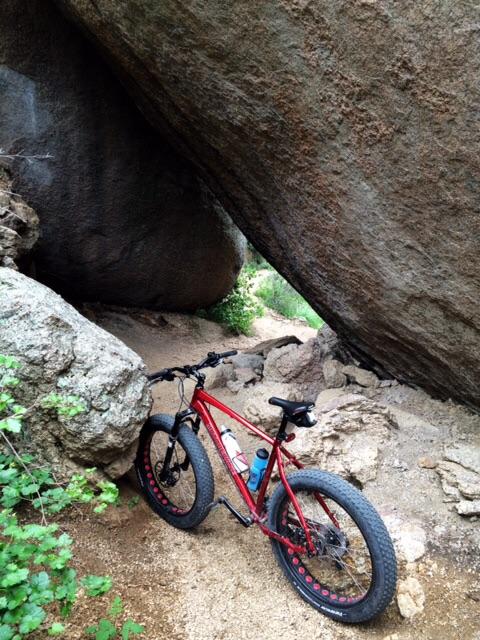 A red fat bike leaning against a large boulder, situated in a rocky outdoor setting surrounded by greenery. The scene captures the rugged terrain and natural beauty of the area. Elk Park Trail mountain bike trail.