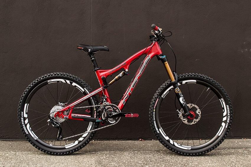 Intense Carbine 275: Mountain bike with a red frame, featuring dual suspension, wide tires, and disc brakes, positioned against a dark wall.