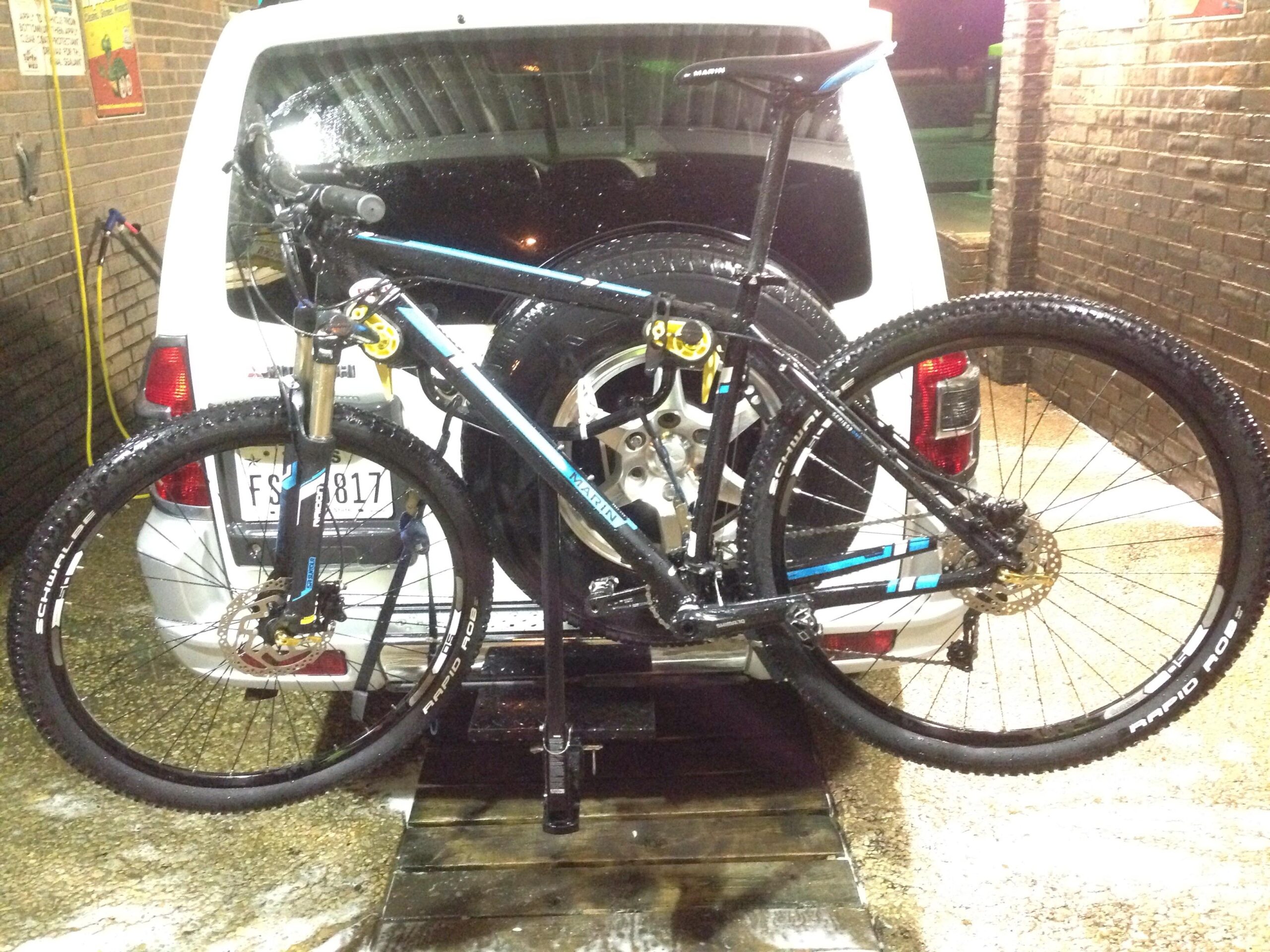 Marin Bobcat Trail: A bicycle being washed at a car wash station, mounted on a bike rack attached to the back of a white vehicle. The bike is partly wet, with water droplets visible, and the surrounding area includes a wooden platform and a brick wall.
